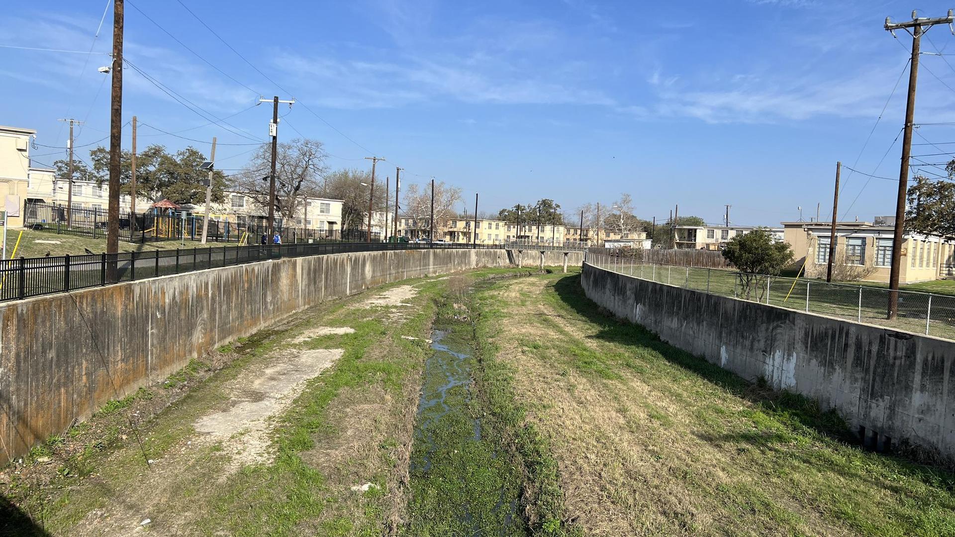 Bright daylight on a concrete channel with sparse water, surrounded by urban buildings.