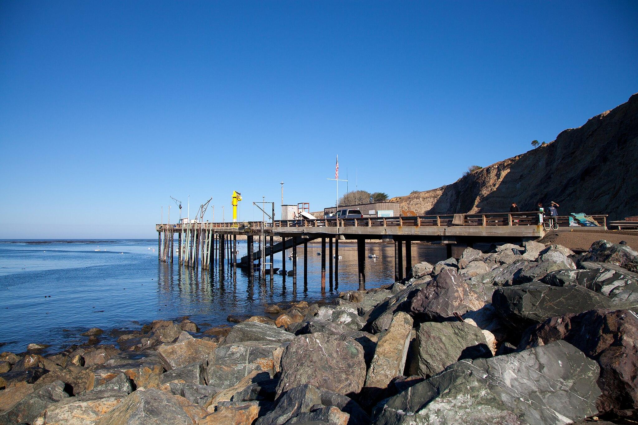 A long wooden pier extends into the ocean from a rocky shore with people.