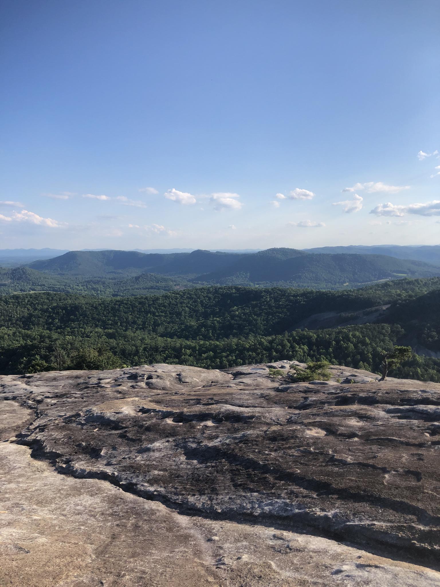 Rocky foreground overlooks distant forested mountains under a clear blue sky.