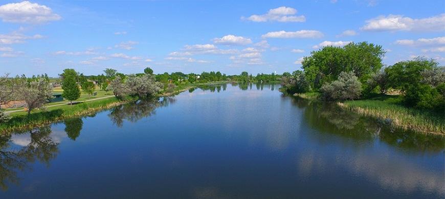 Wide view of a calm lake reflecting blue sky and green trees on a sunny day.