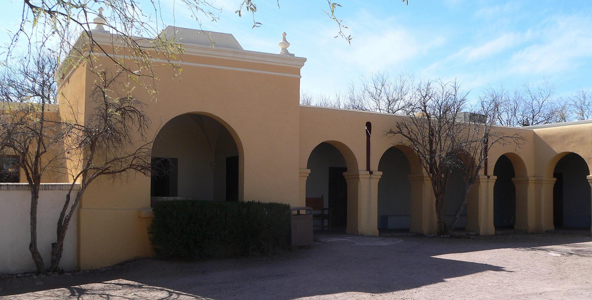 Yellow adobe-style building with arched walkways and trees under a partly cloudy sky.