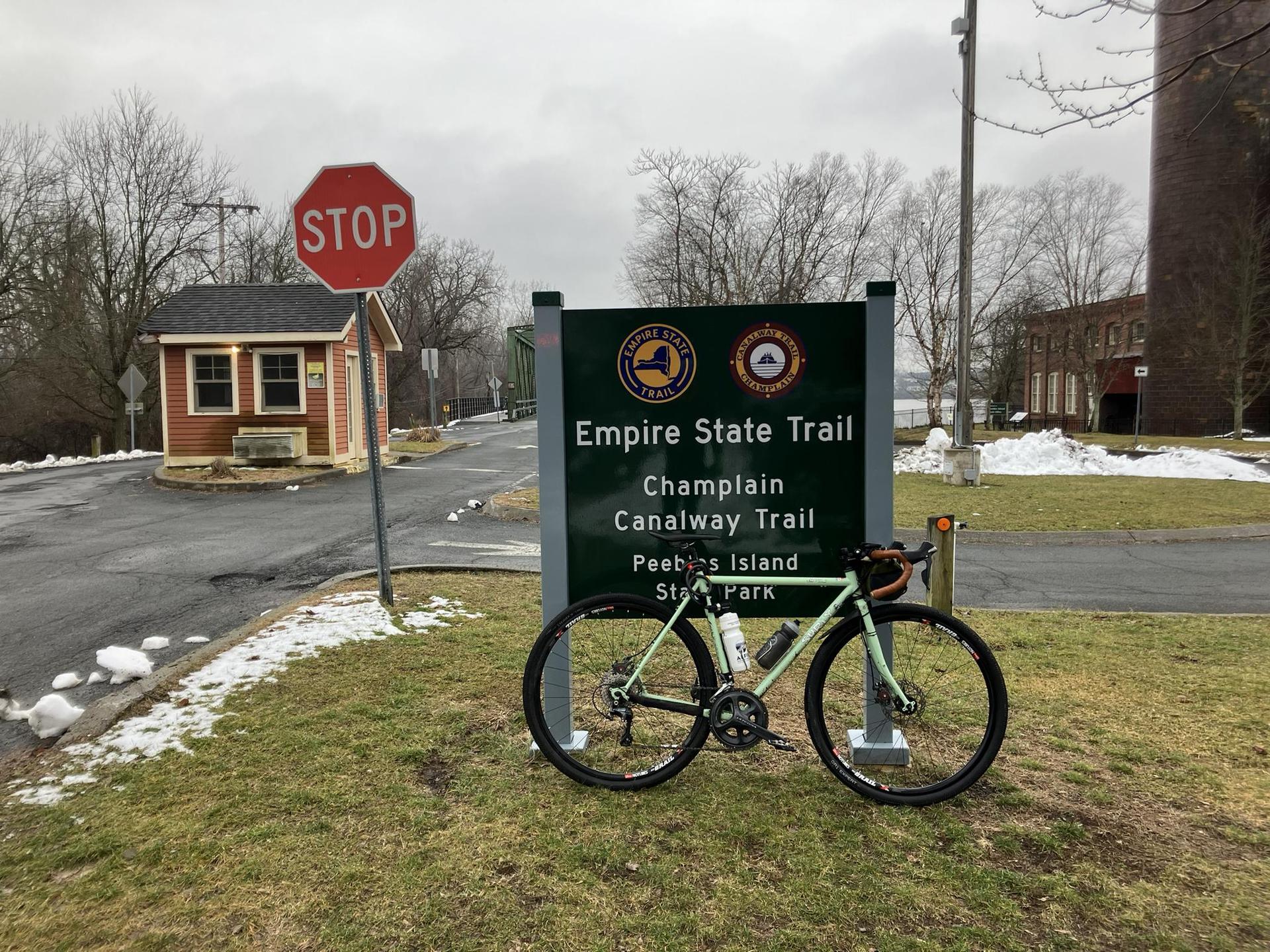 A bicycle leans against a trail sign on a cloudy day with snow patches.