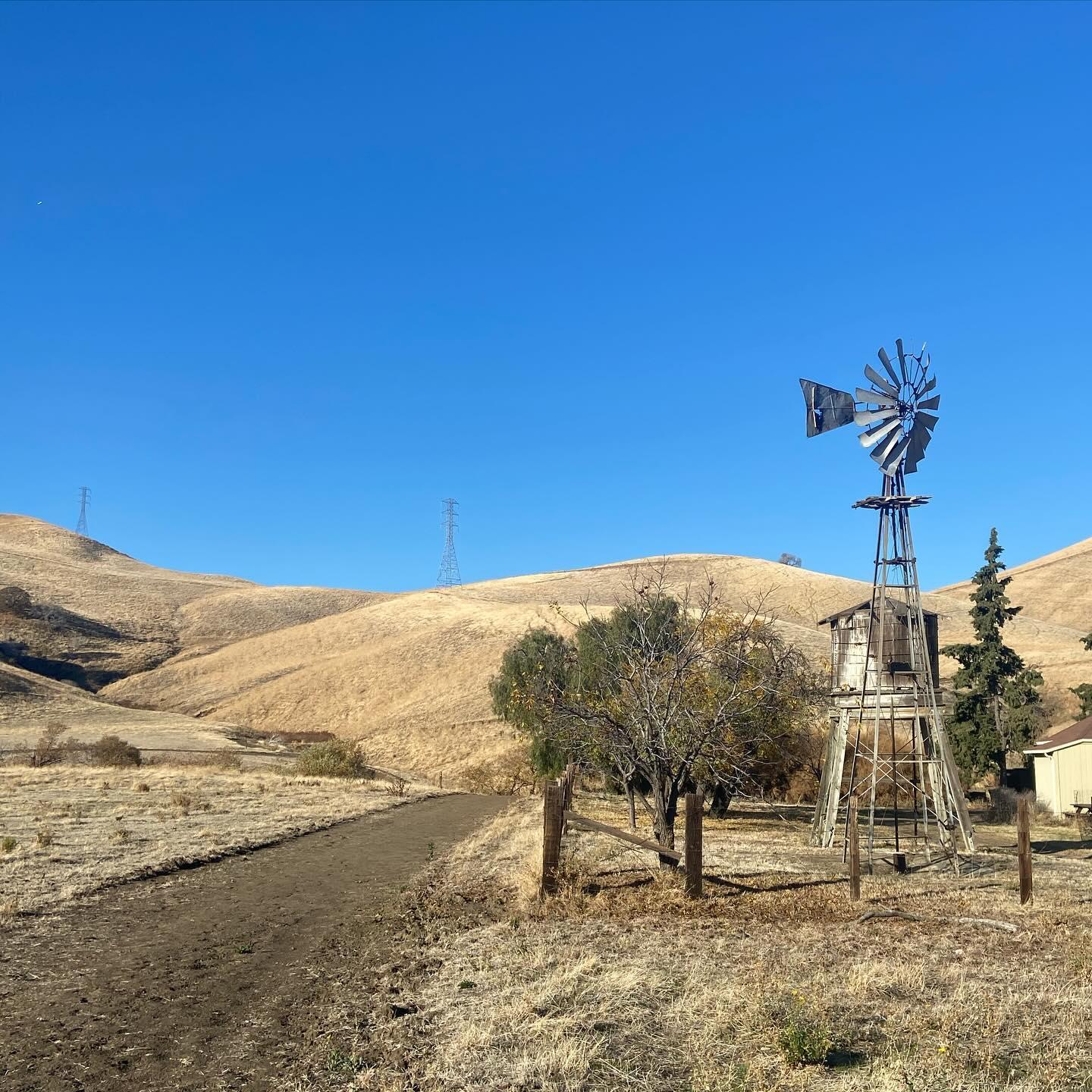 A rustic windmill stands beside a dirt path in sunny, dry rolling hills.