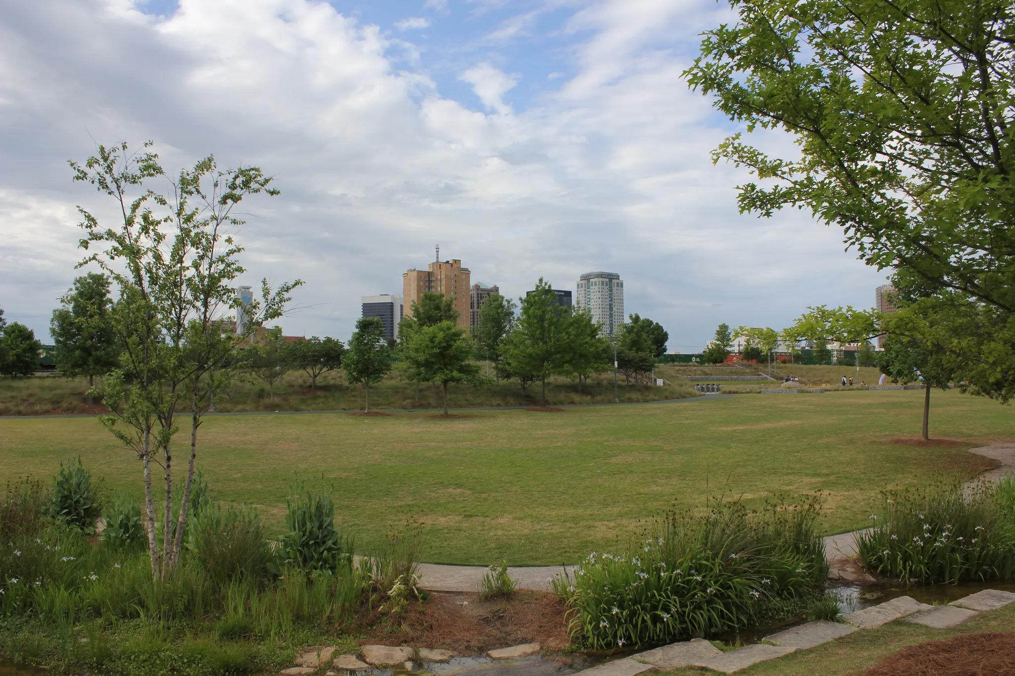 Expansive green park with trees, distant city skyline, and people under cloudy sky. - Railroad Park – Railroad Park loop from Avondale