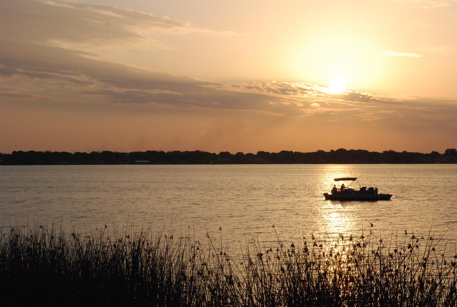 A boat with people on a calm lake at golden sunset, reeds in foreground.