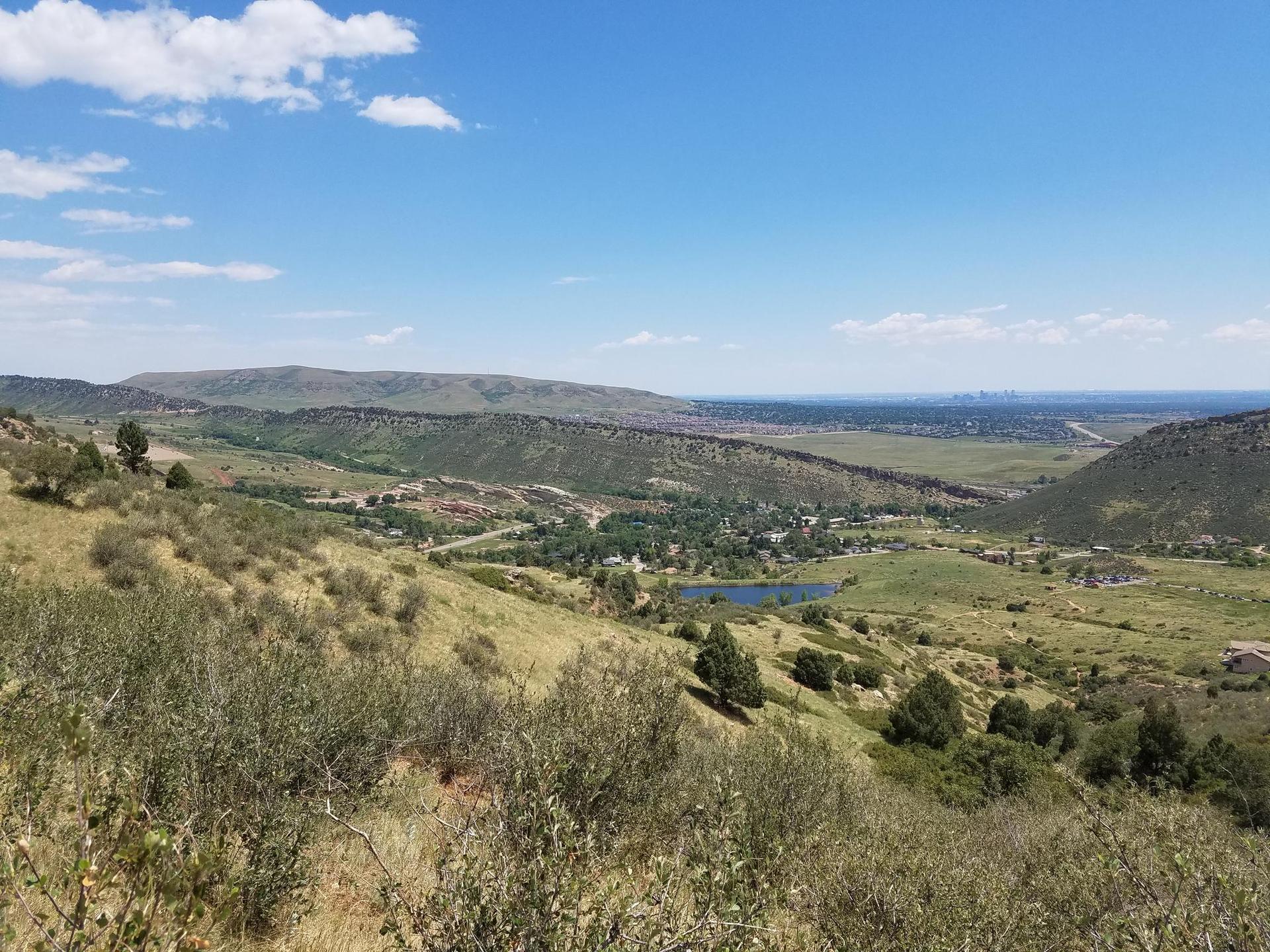 Expansive valley view with a lake, green hills, and distant structures under blue sky.