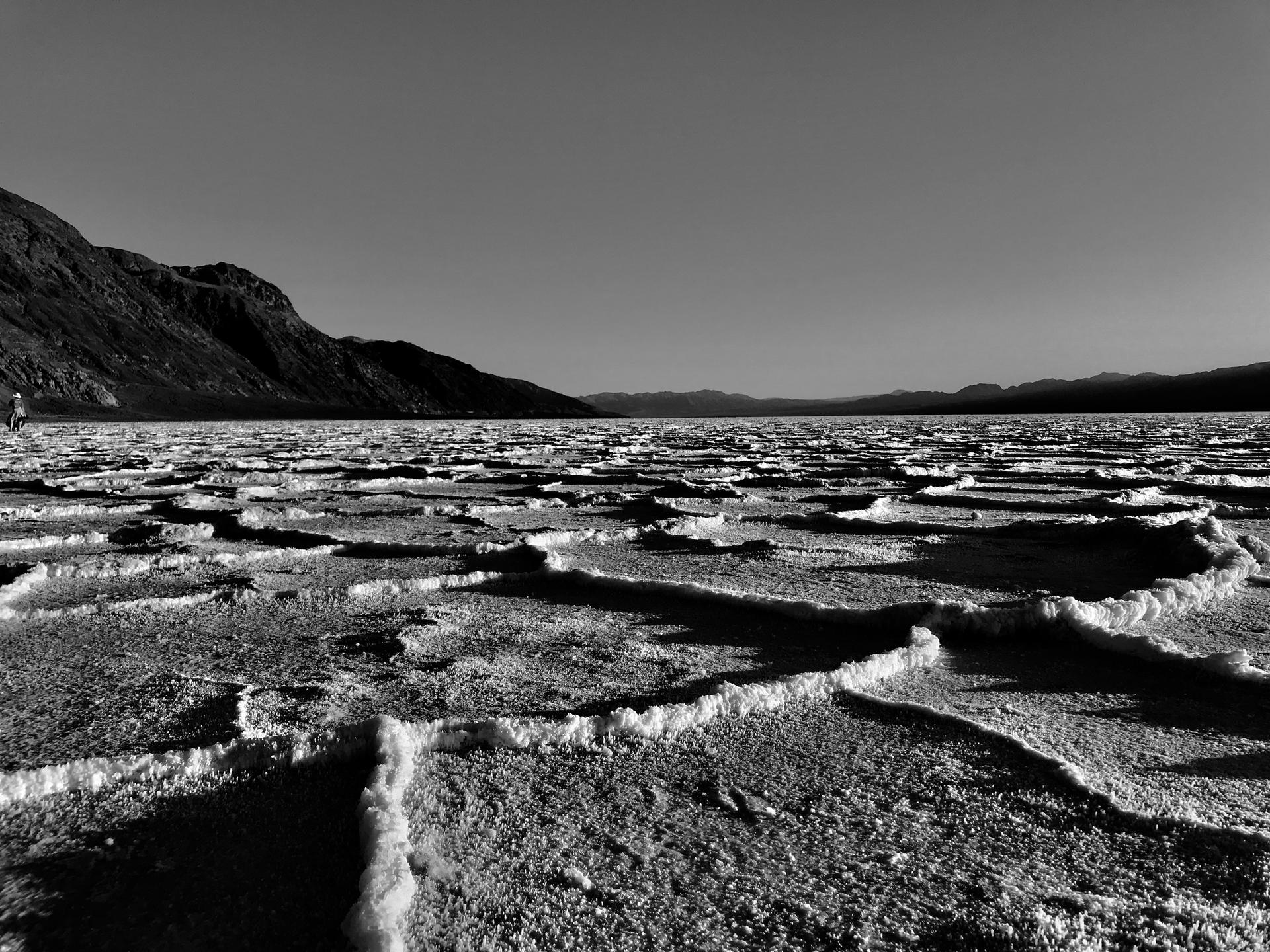 Black and white view of textured salt formations on a vast flat, distant mountains.