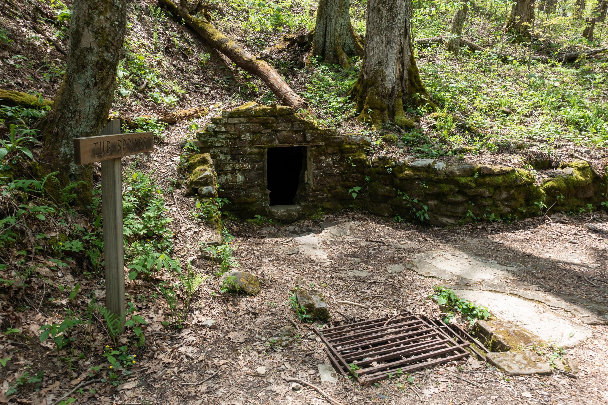 A stone structure and wooden sign in a sun-dappled forest clearing.