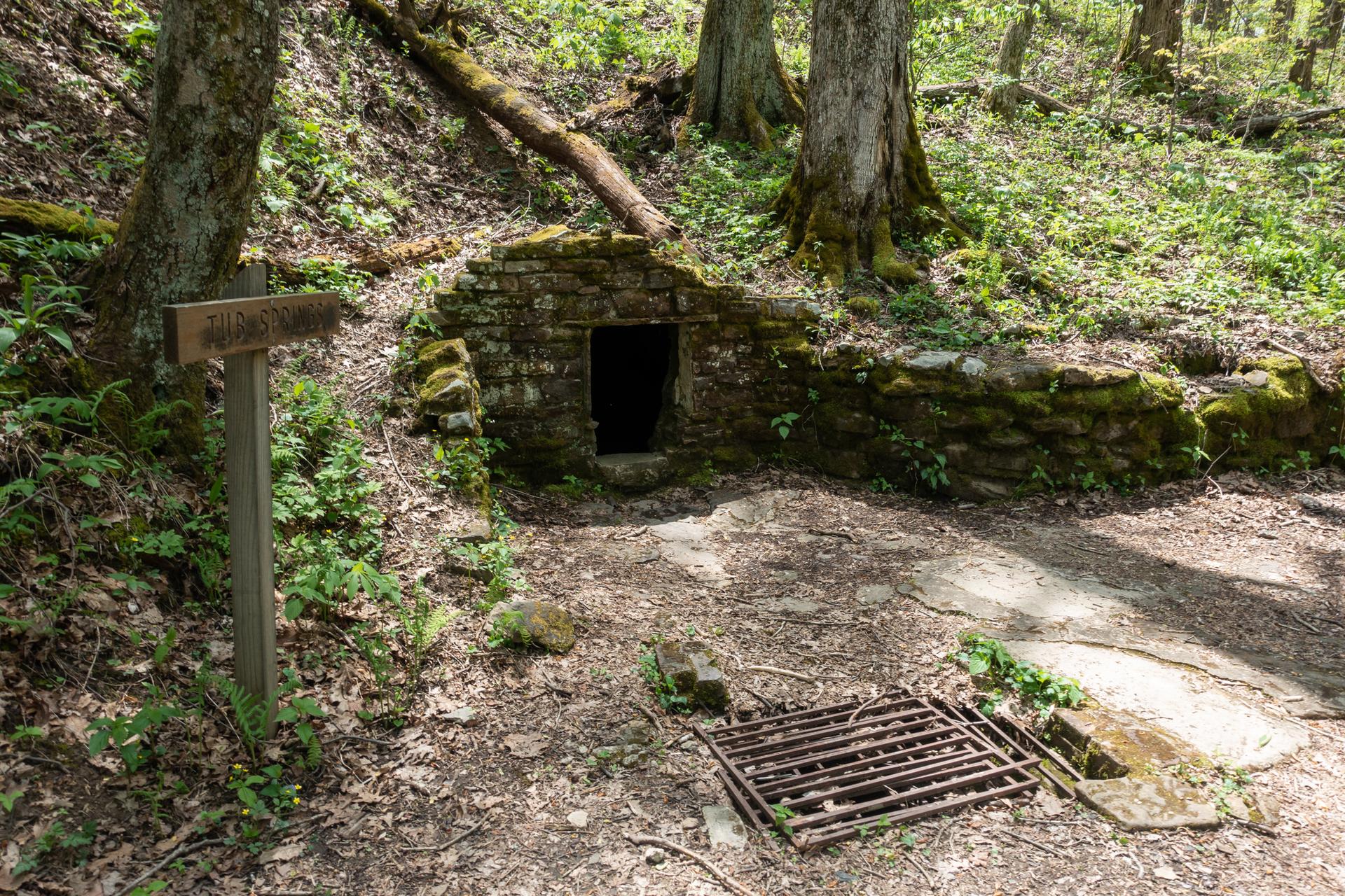 A stone structure and wooden sign in a sun-dappled forest clearing.