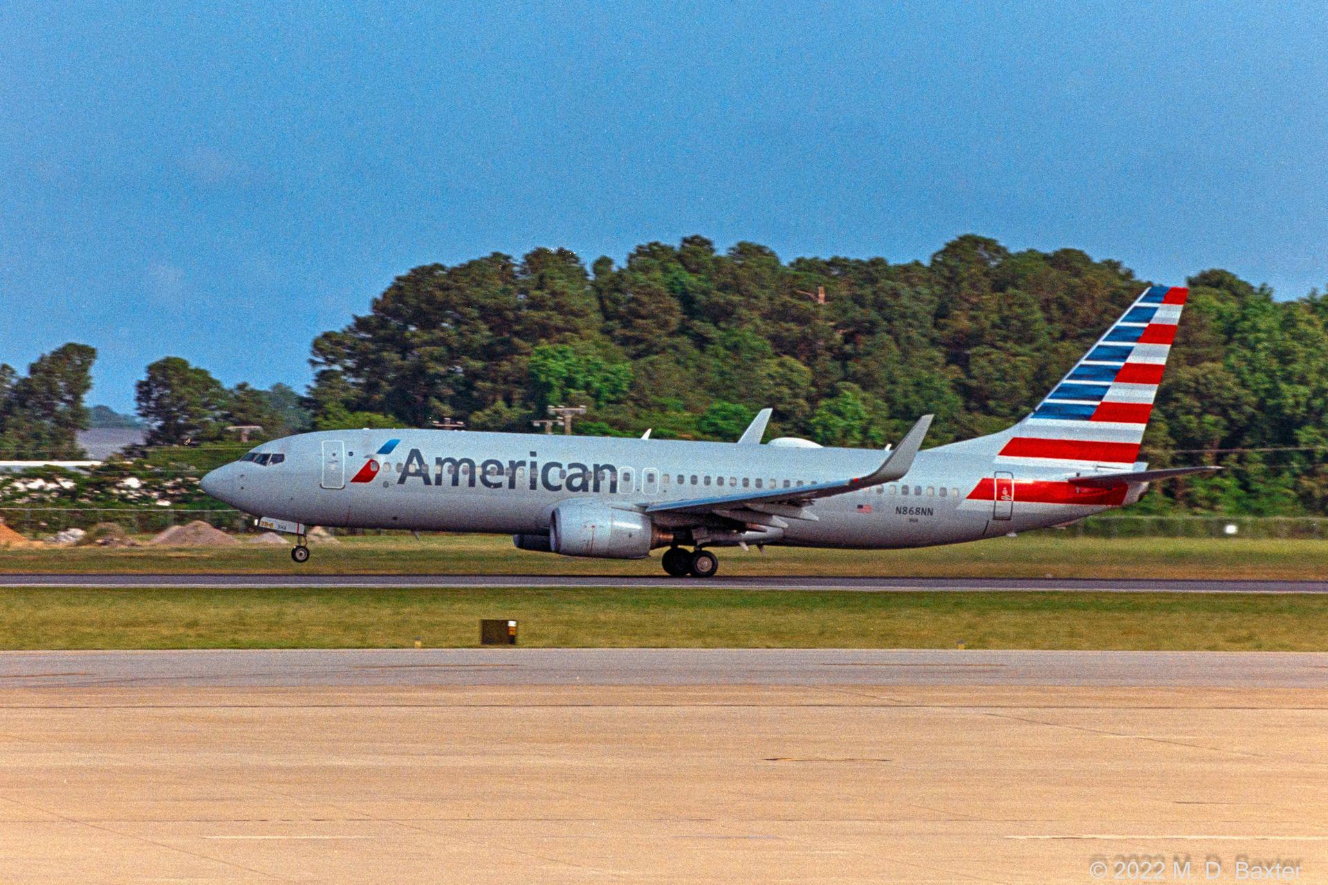 An American Airlines plane on a runway with green trees in daylight.