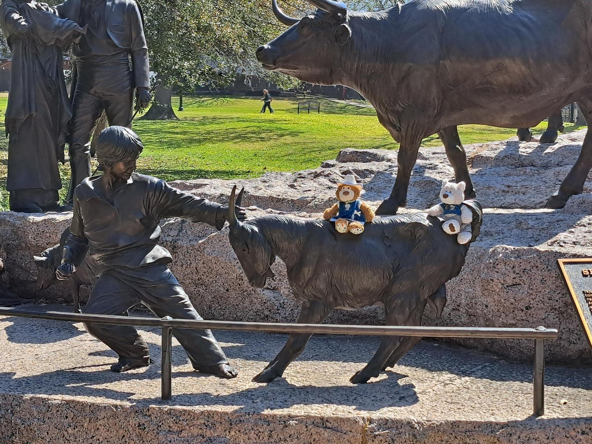 Bronze statues of people, a bull, and a goat with stuffed animals on a sunny day.