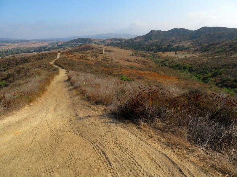 A wide dirt trail winds through dry, rolling hills under a clear sky.