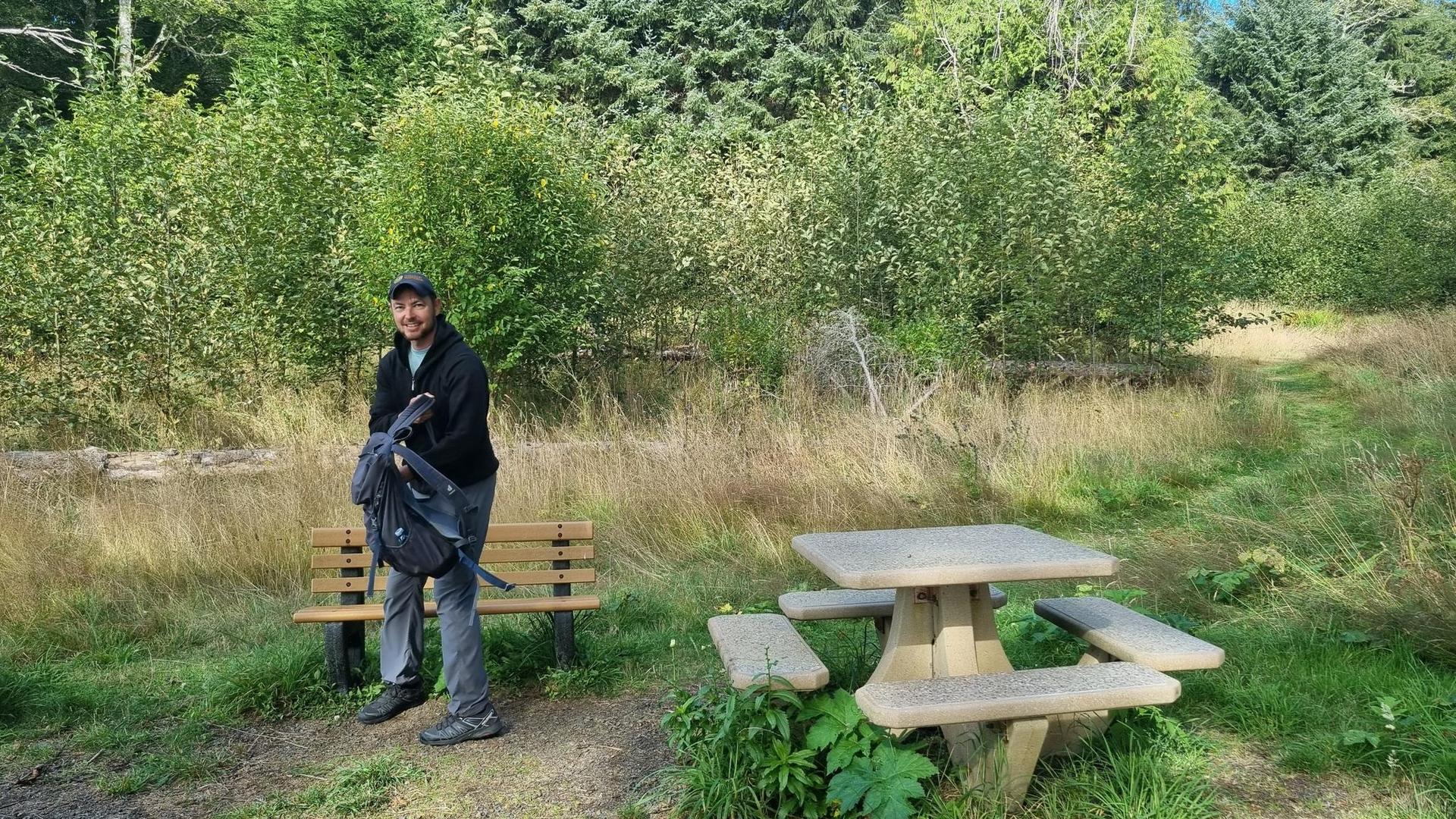 Man with backpack stands by a wooden picnic table in a sunny clearing.