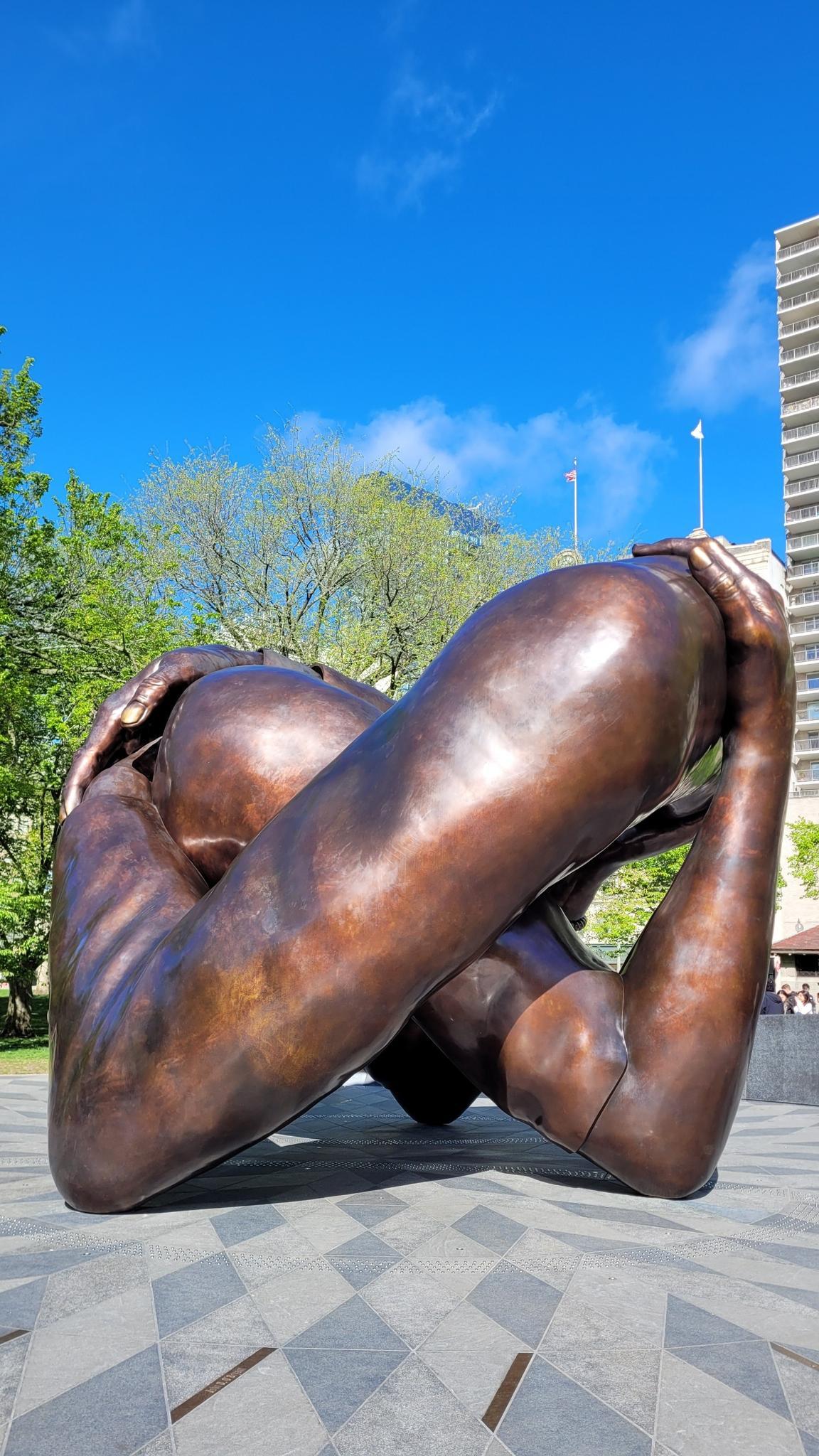 Large bronze sculpture of a person curled up on patterned pavement, blue sky.