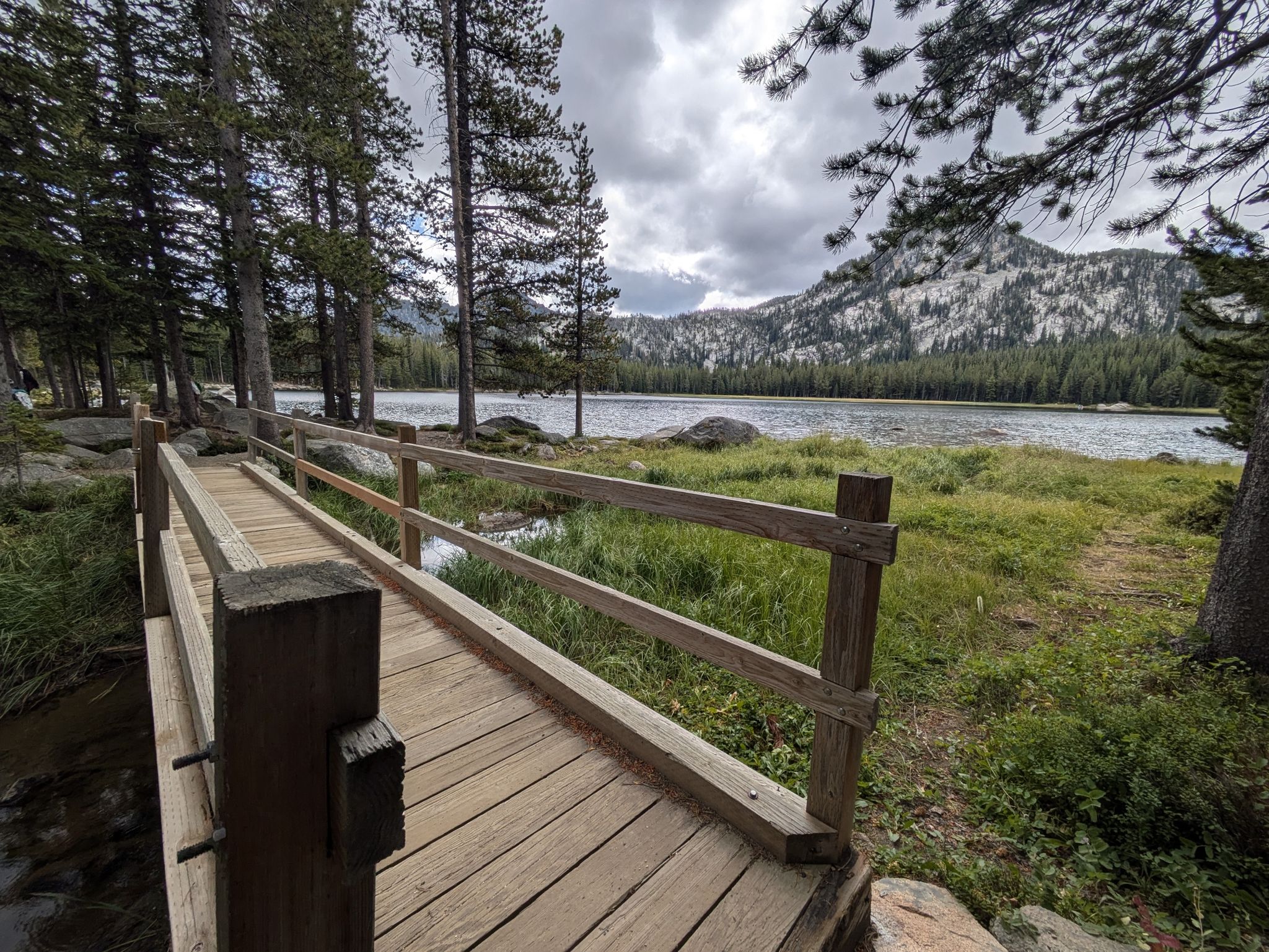 Wooden bridge leads to a grassy path beside a lake and pine forest. - Elkhorn Crest, Crawfish Basin, and Hoffer Lakes Loop via Anthony Lake