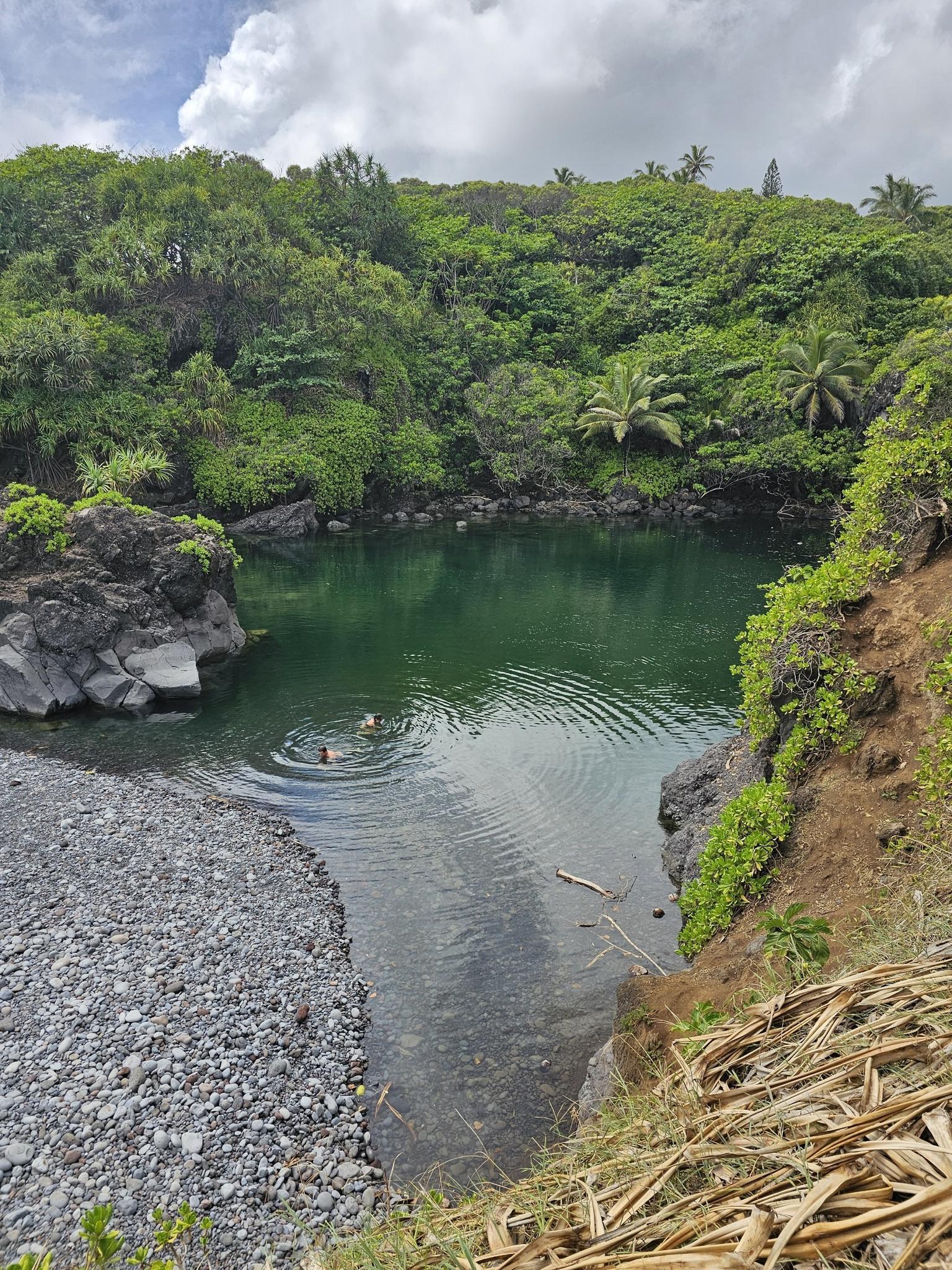 Two people swimming in a green-blue natural pool surrounded by lush hills.