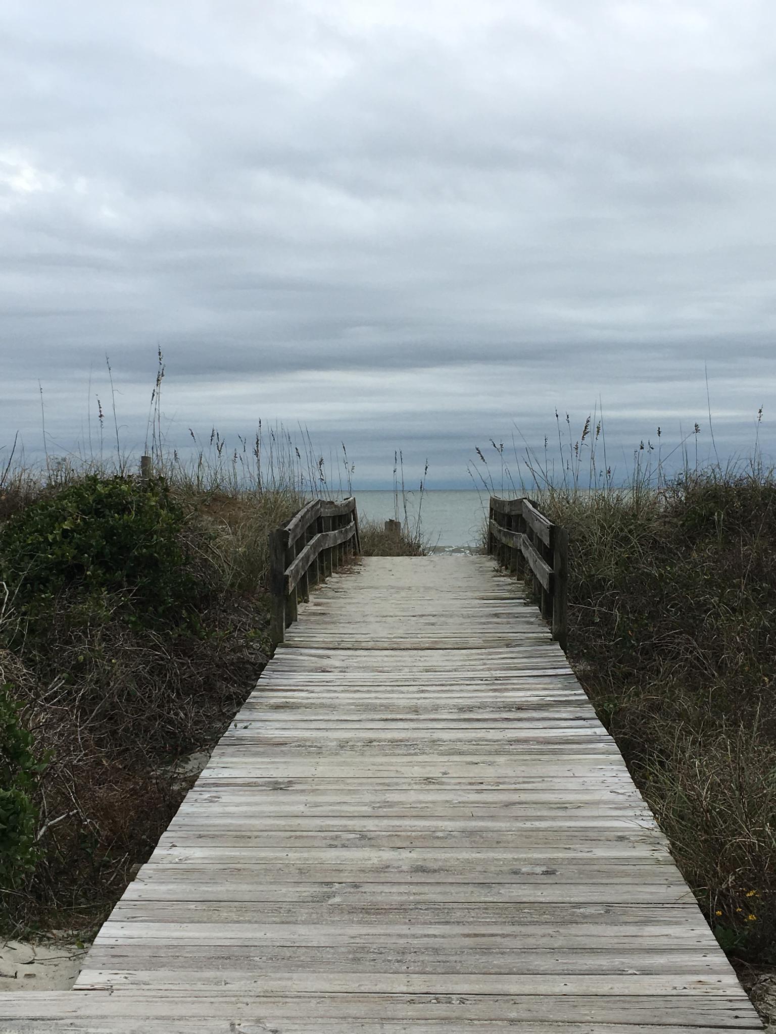 Wooden boardwalk leads through dunes to the ocean under an overcast sky.