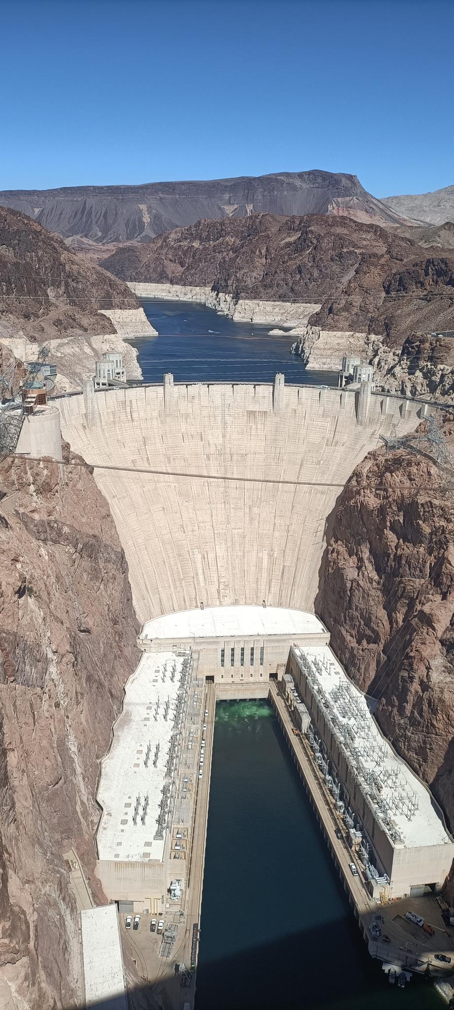 Massive concrete dam in a deep canyon with a river and clear blue sky.