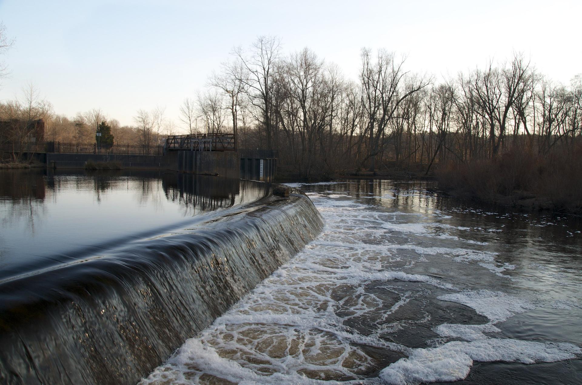 Water flows over a dam into a foamy river, surrounded by bare trees.