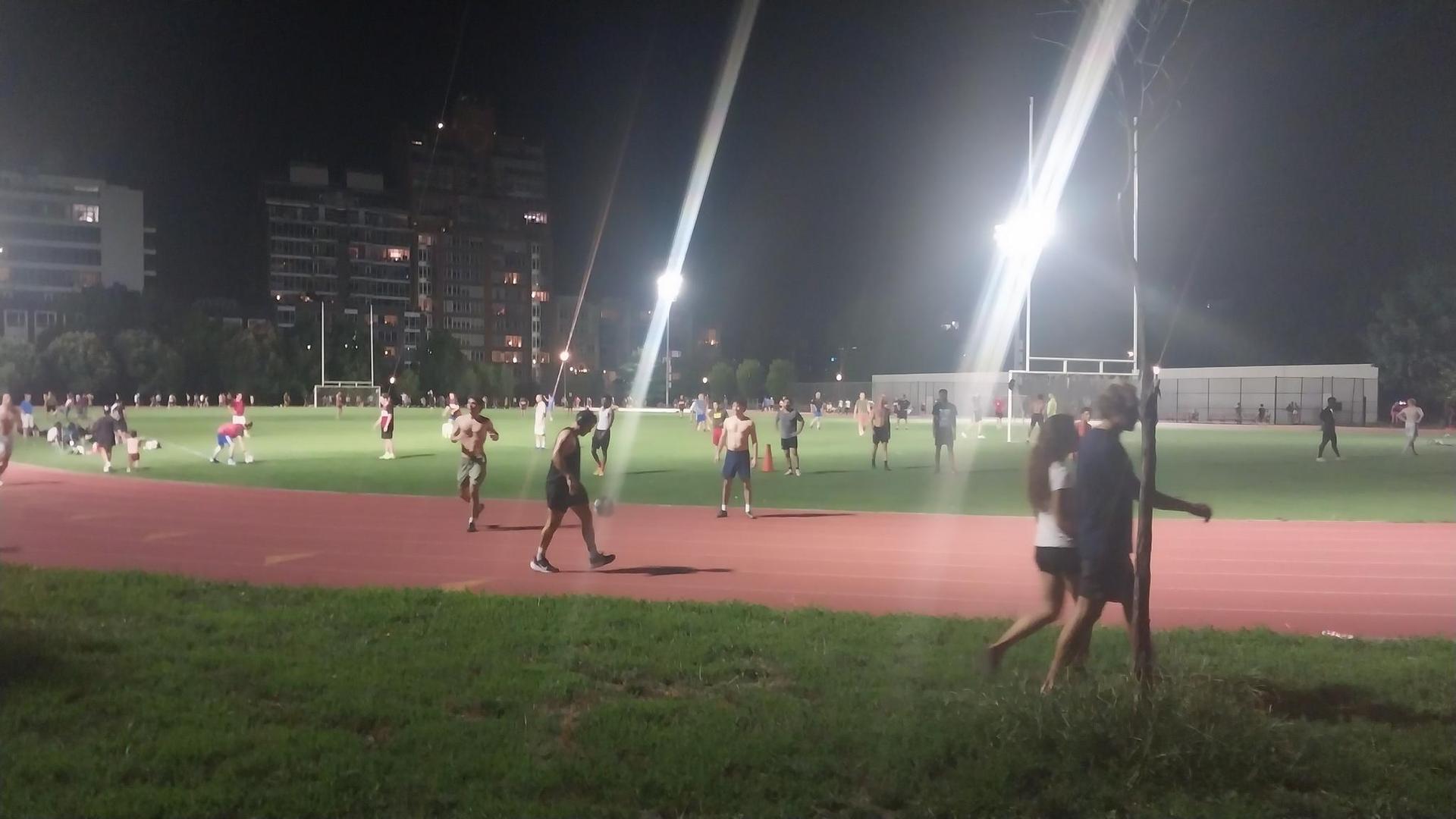 People walk and run on a brightly lit sports field at night.