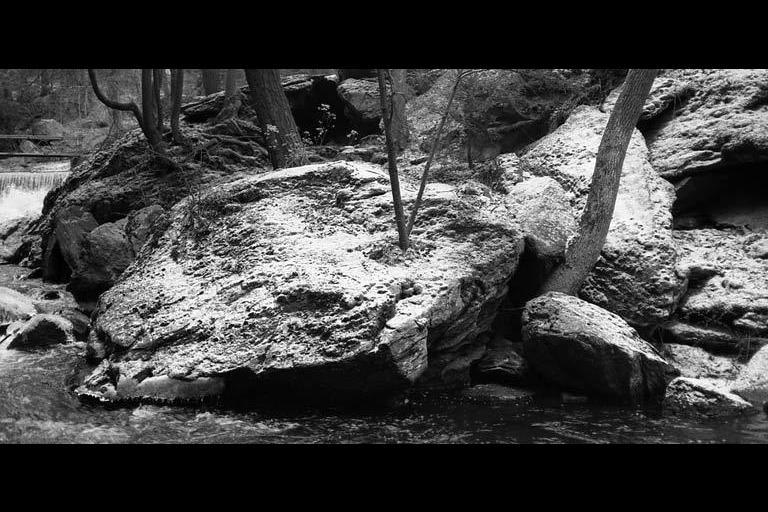 Black and white photo of large rocks and trees in a flowing stream.