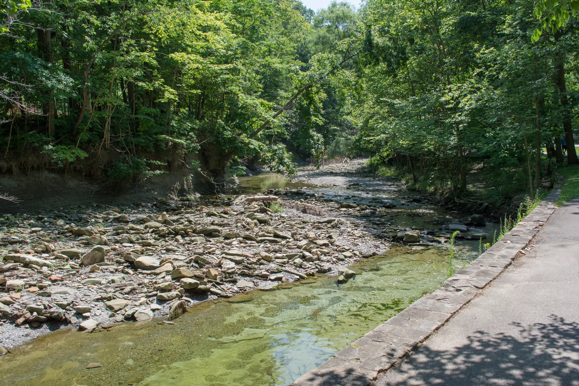 Sunny view of a shallow, rocky riverbed next to a paved path and forest.