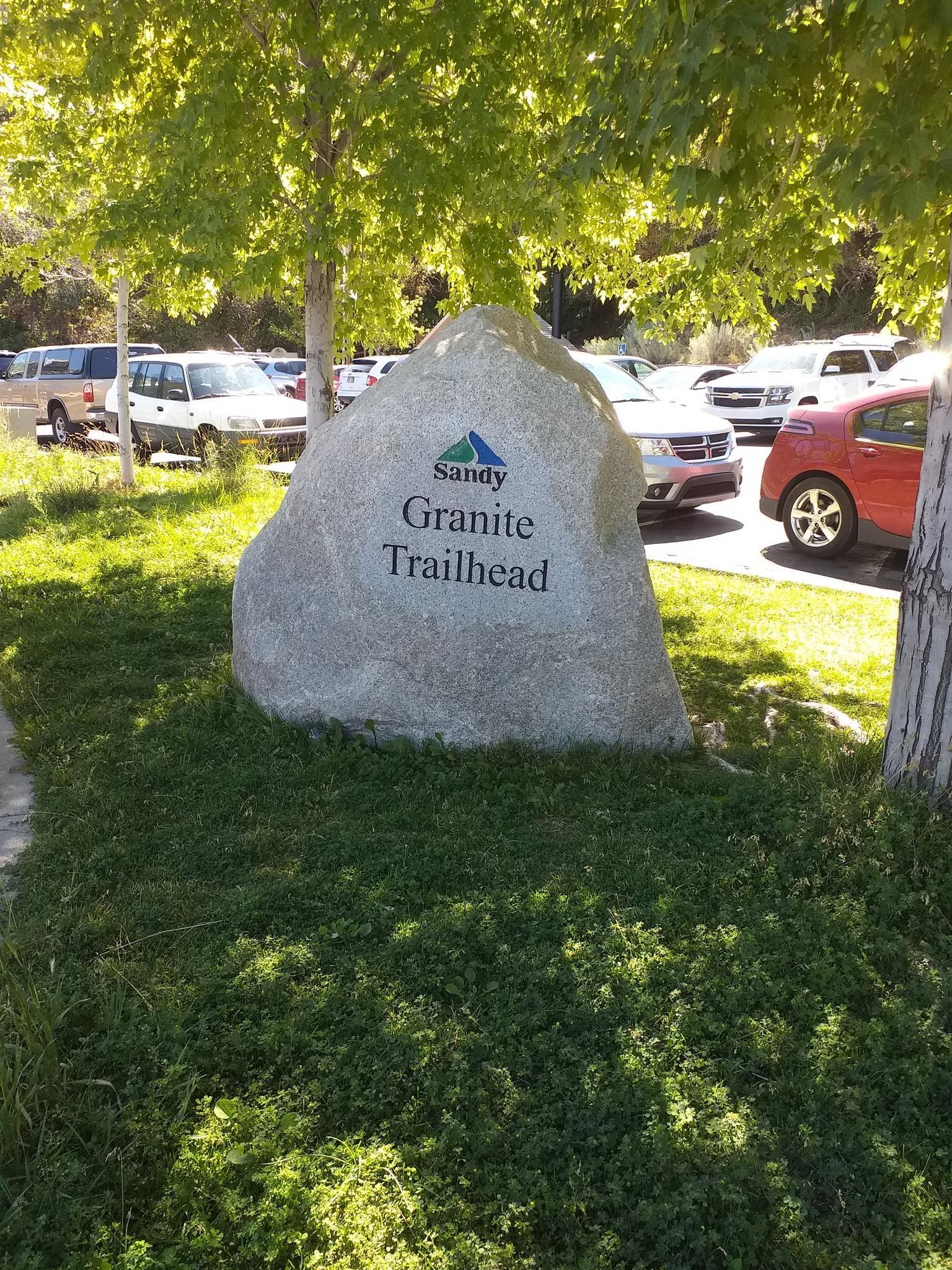 A large rock sign in a grassy area with cars and trees.