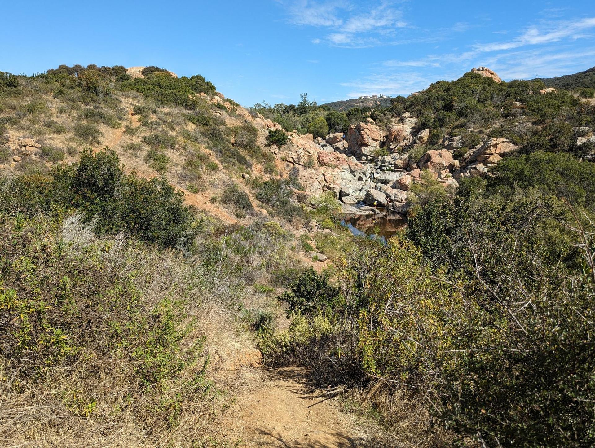 A dirt path leads through brushy hills towards a rocky canyon and water.
