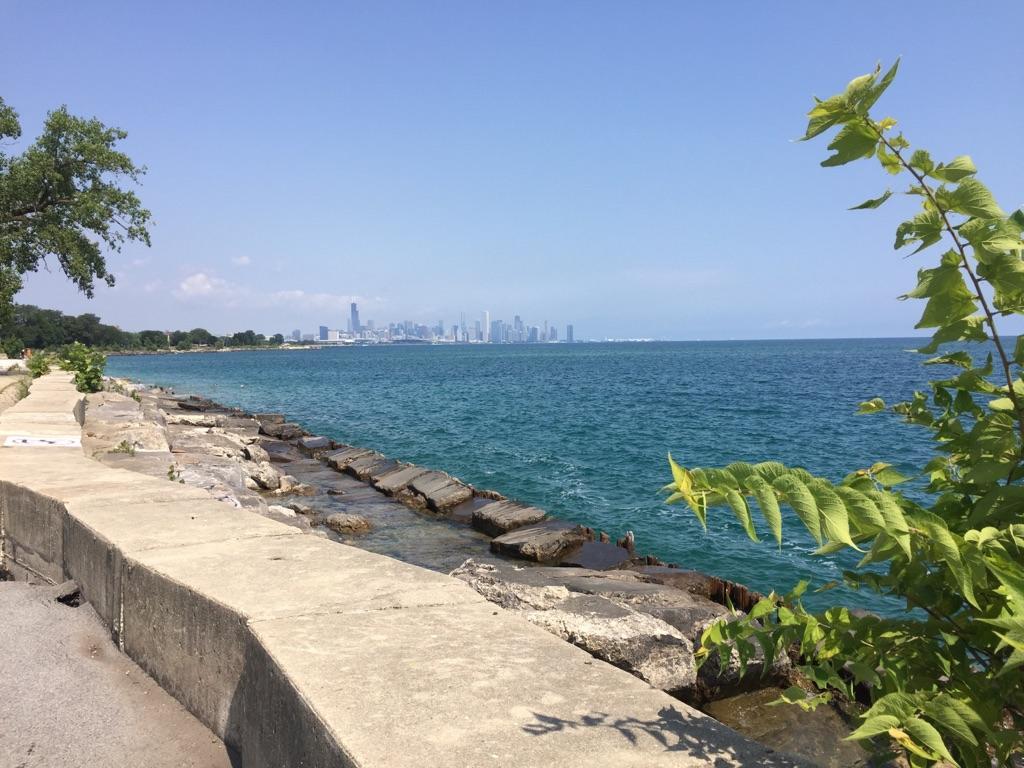 Rocky shoreline next to blue water, distant city skyline under clear sky.