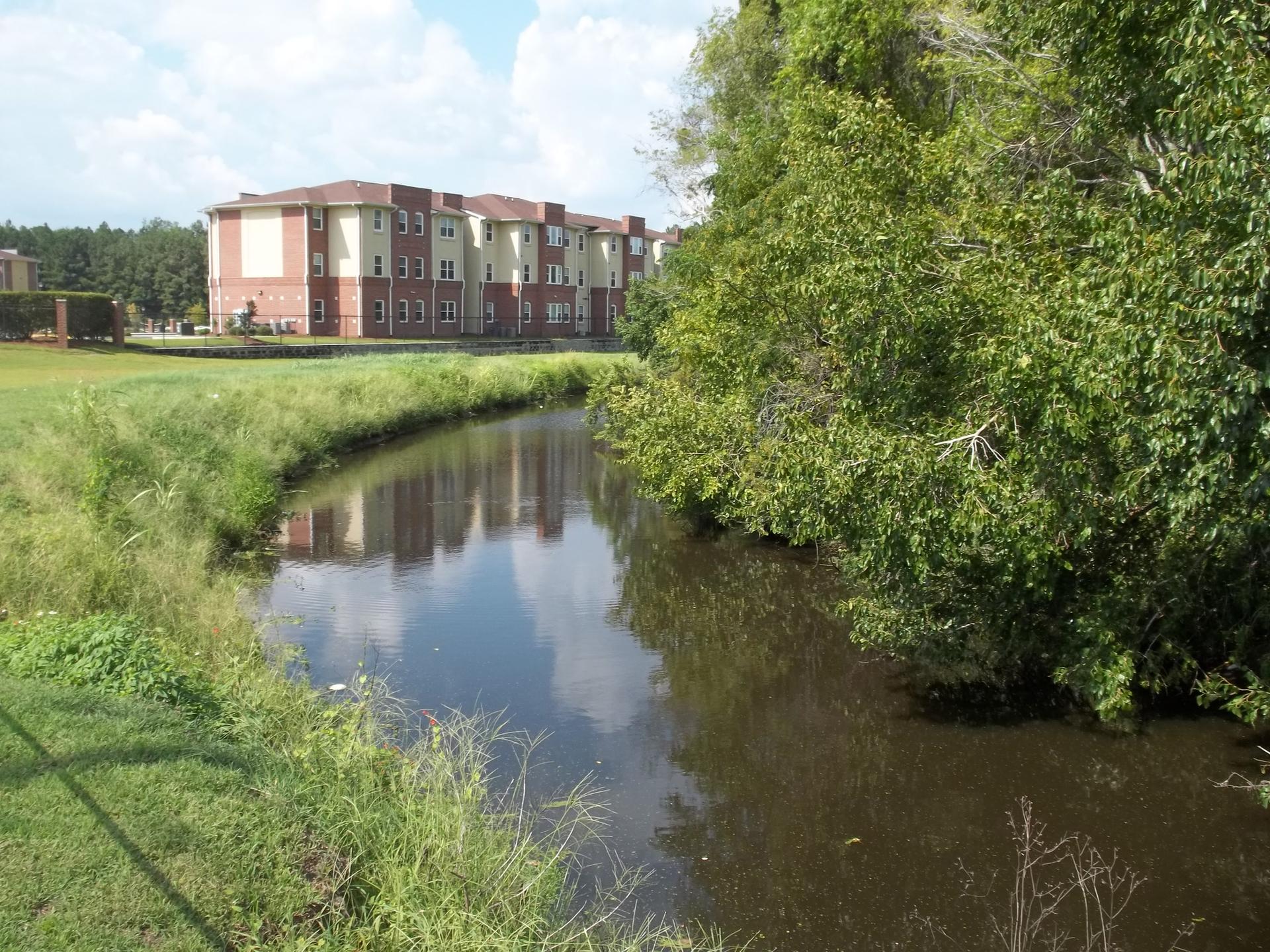 A calm river flows past green banks and distant brick buildings.