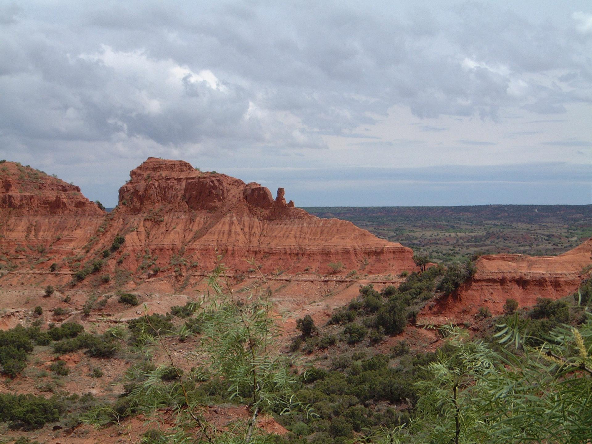Rugged red rock formations and green brush under a cloudy sky.