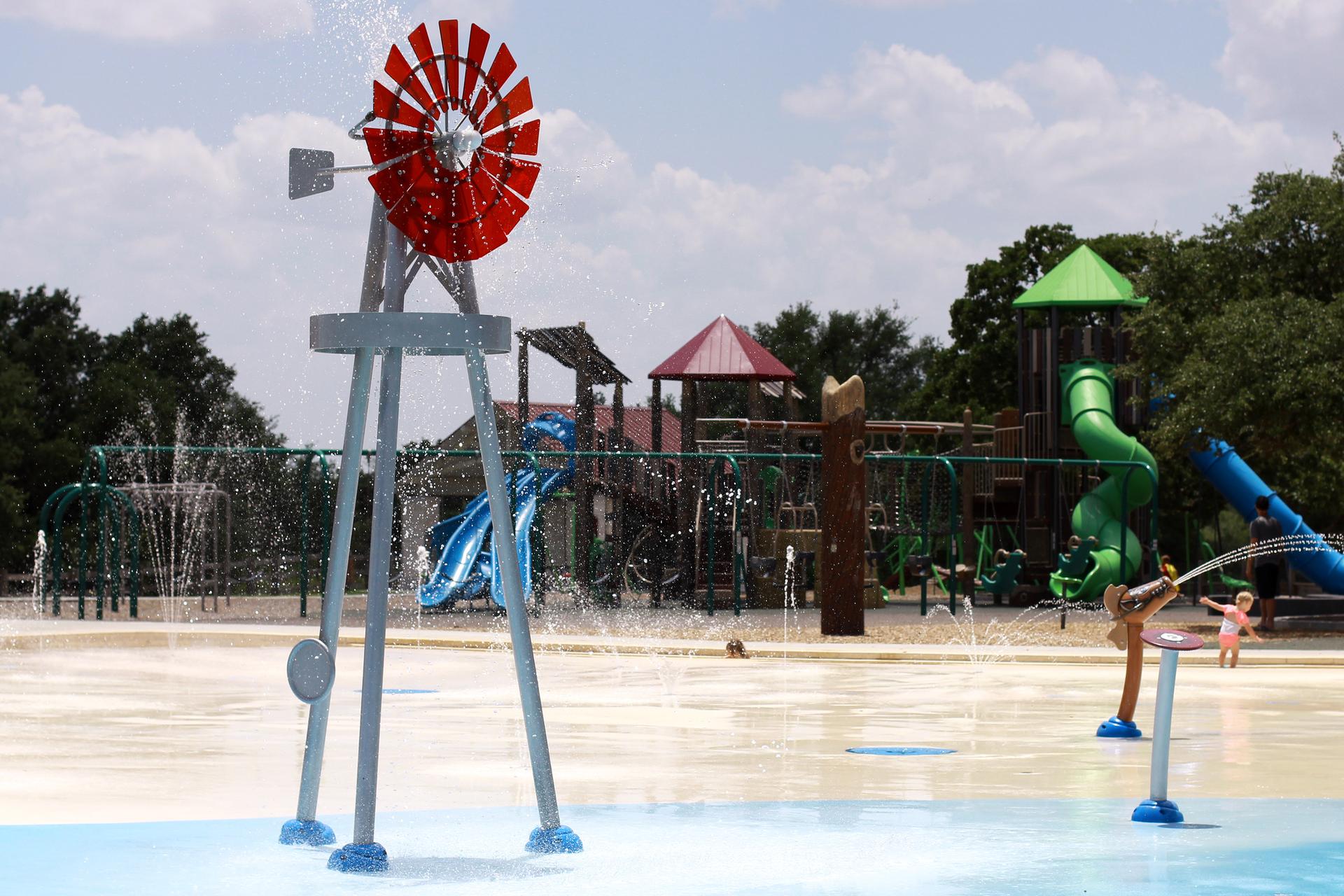 Kids play on a sunny splash pad with a red windmill and slides.