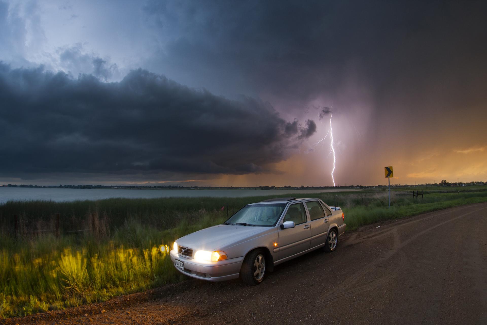 Silver car on dirt road under dramatic stormy sky with lightning strike.