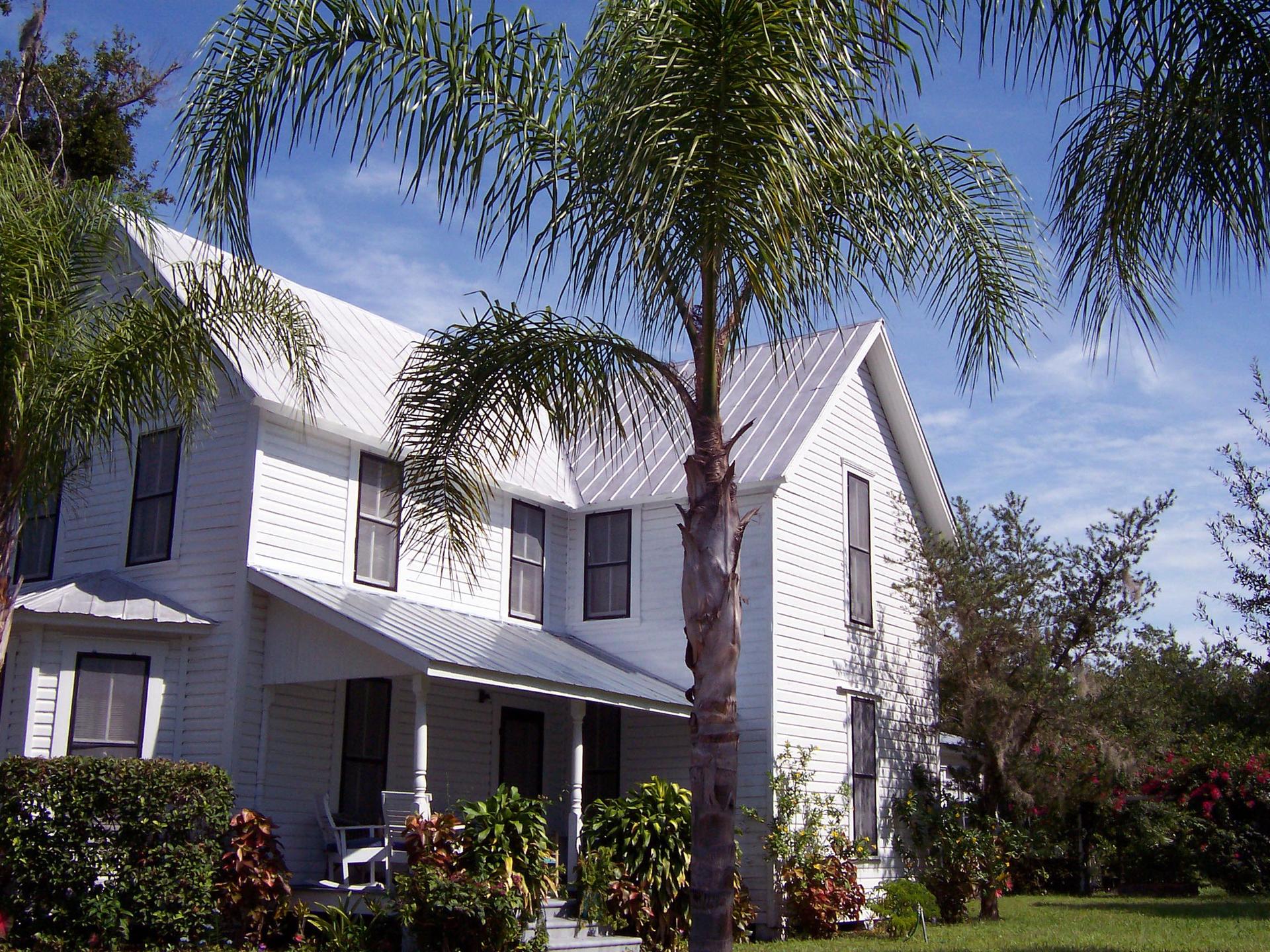 White two-story house with metal roof, surrounded by palm trees on a sunny day.