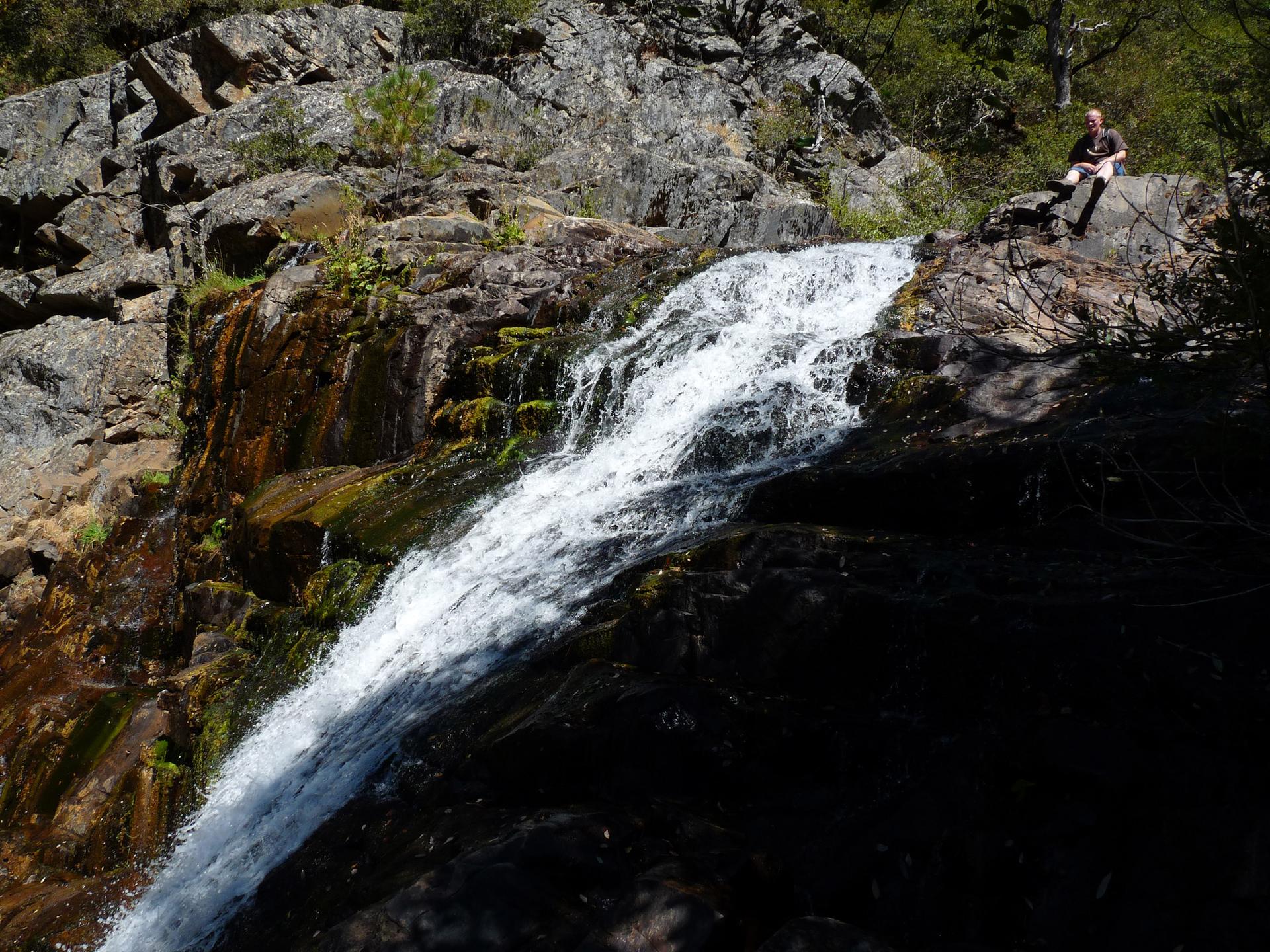 Person sitting on rocks beside a cascading waterfall in bright sunlight.