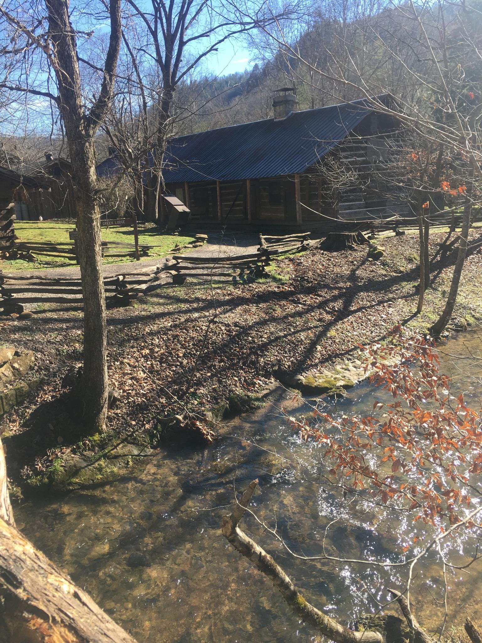 A rustic log cabin sits beside a clear stream in a sunny, bare winter forest.