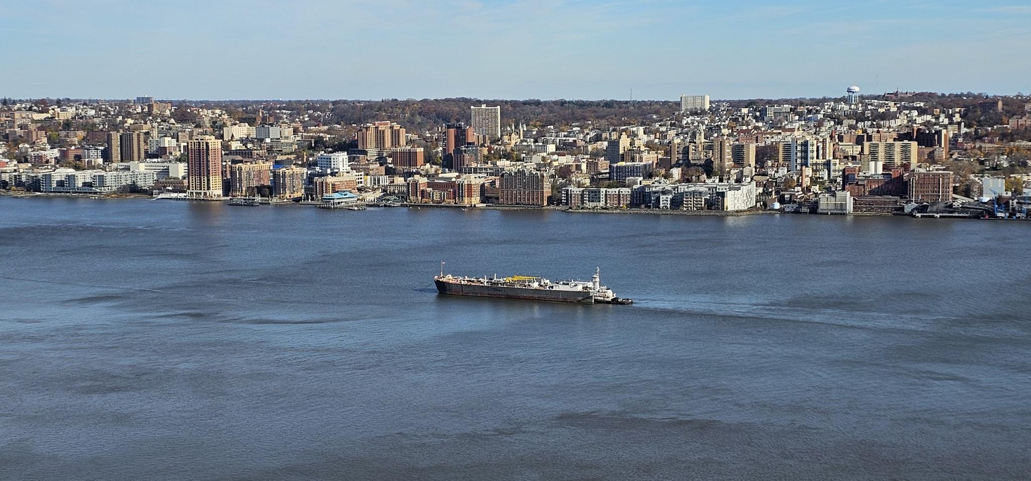 Large cargo ship on wide river with bustling city skyline under clear sky.