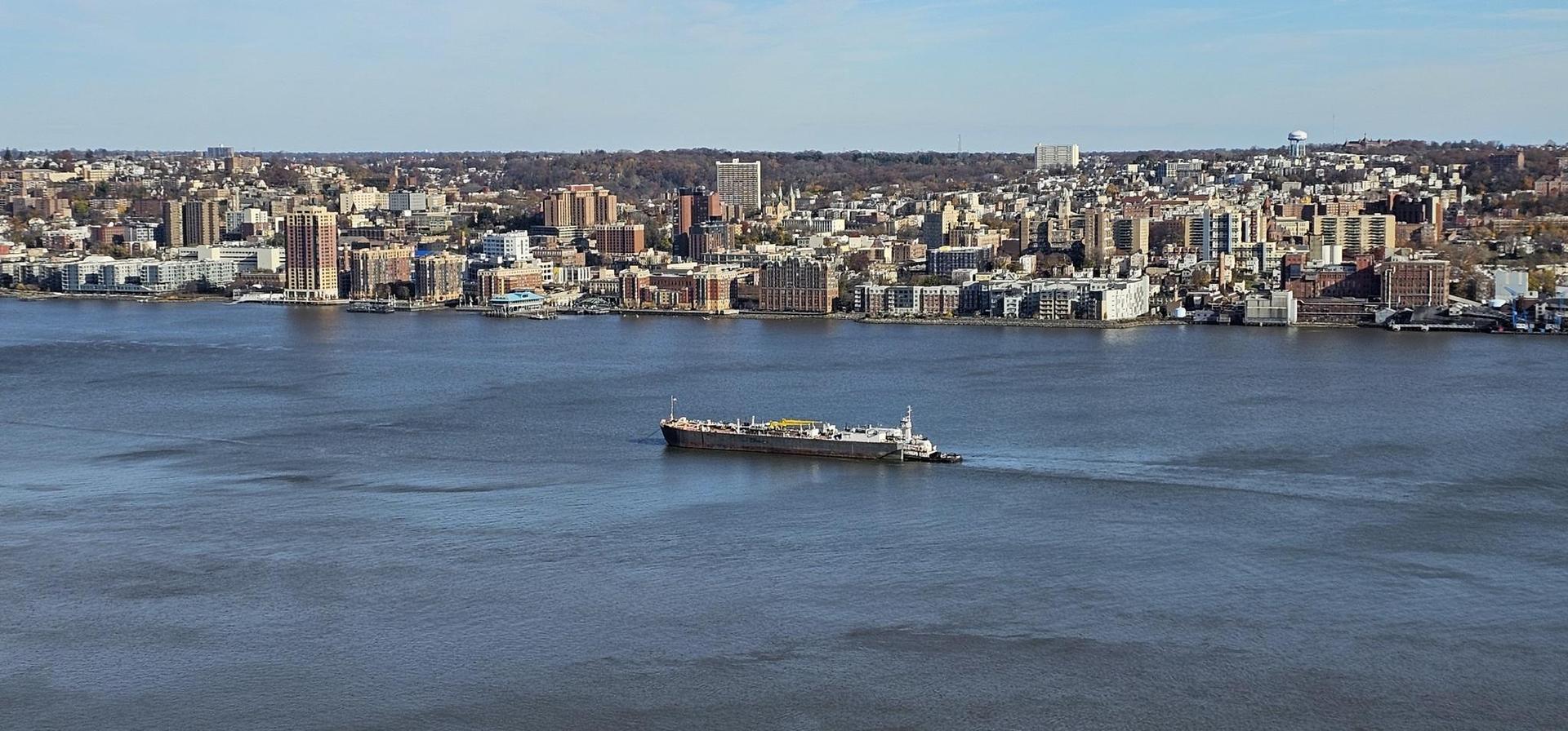 Large cargo ship on wide river with bustling city skyline under clear sky.