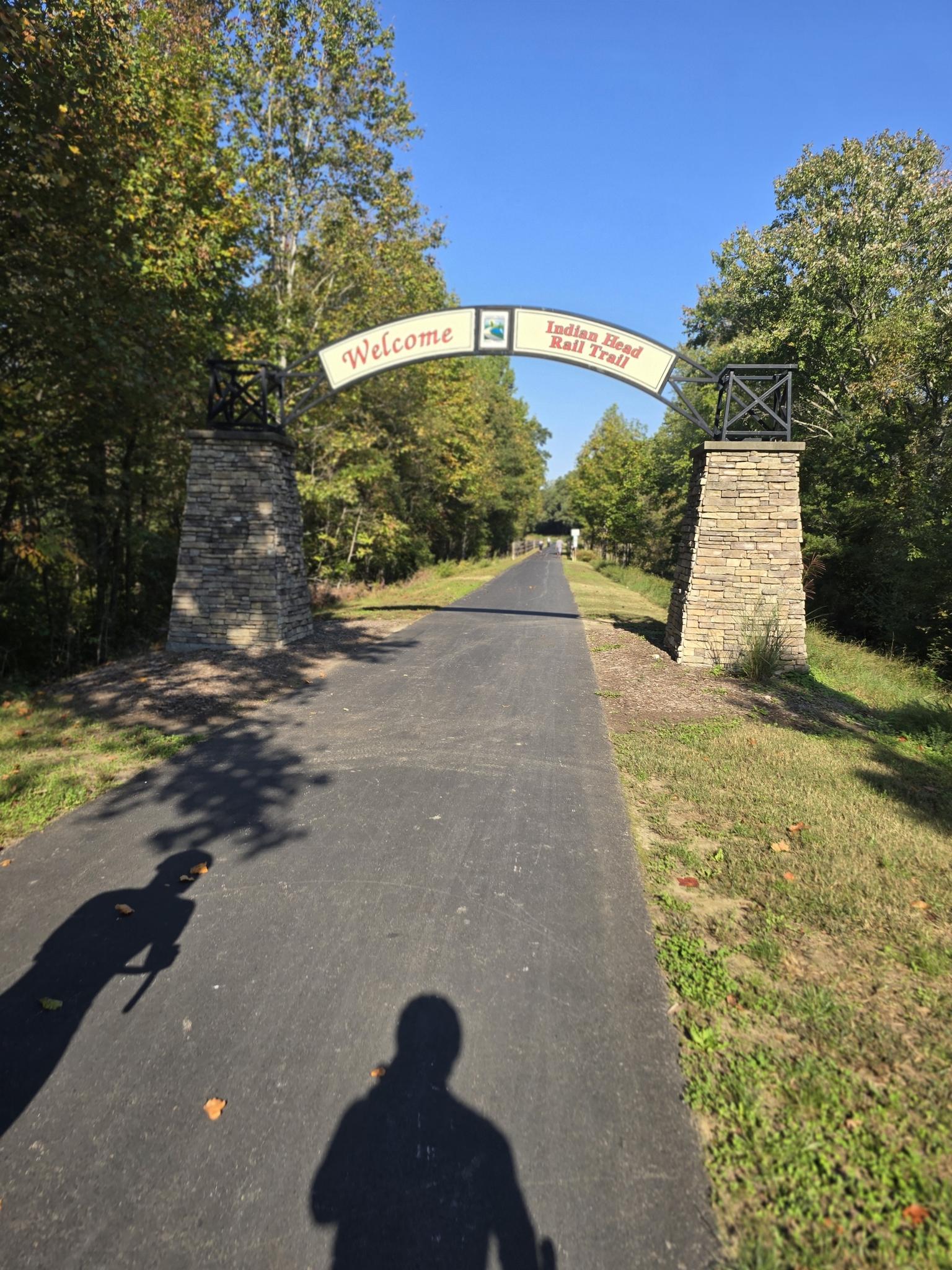 Paved path through a "Welcome" archway, lined with trees on a sunny day.