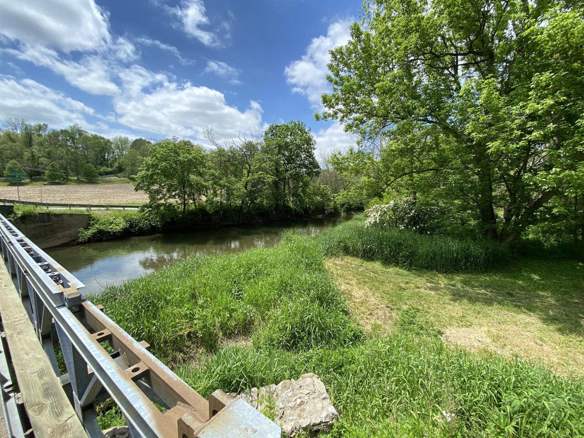 A river flows past a grassy bank, seen from a bridge on a sunny day.