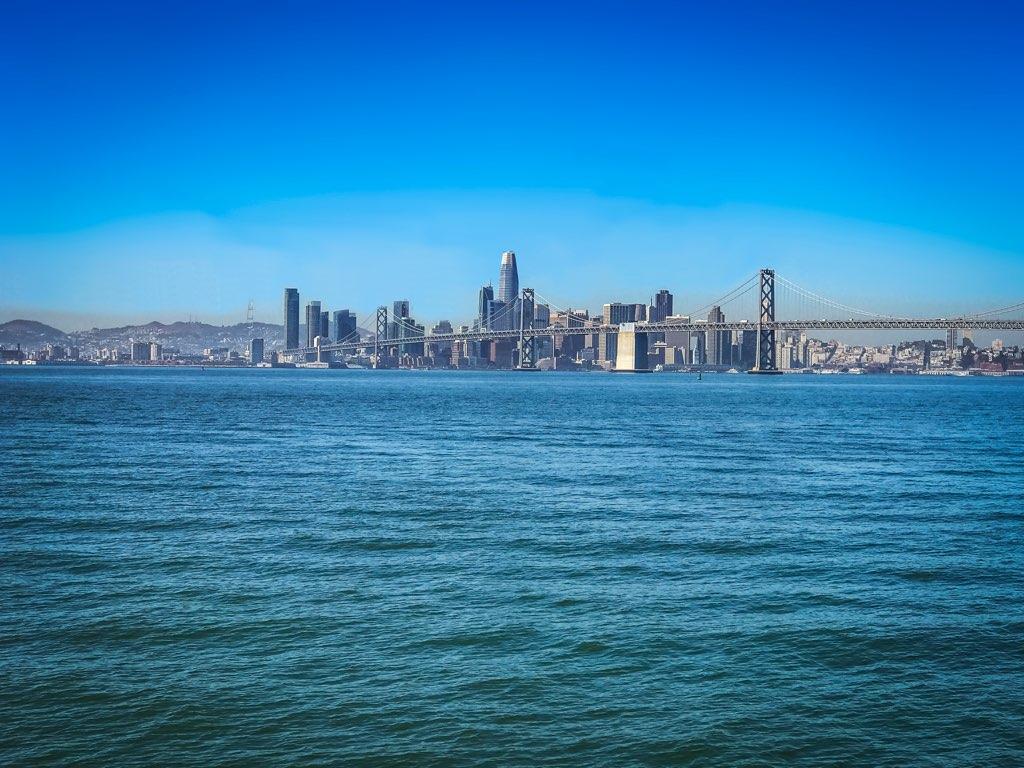 City skyline and bridge across a wide body of water under clear blue sky.