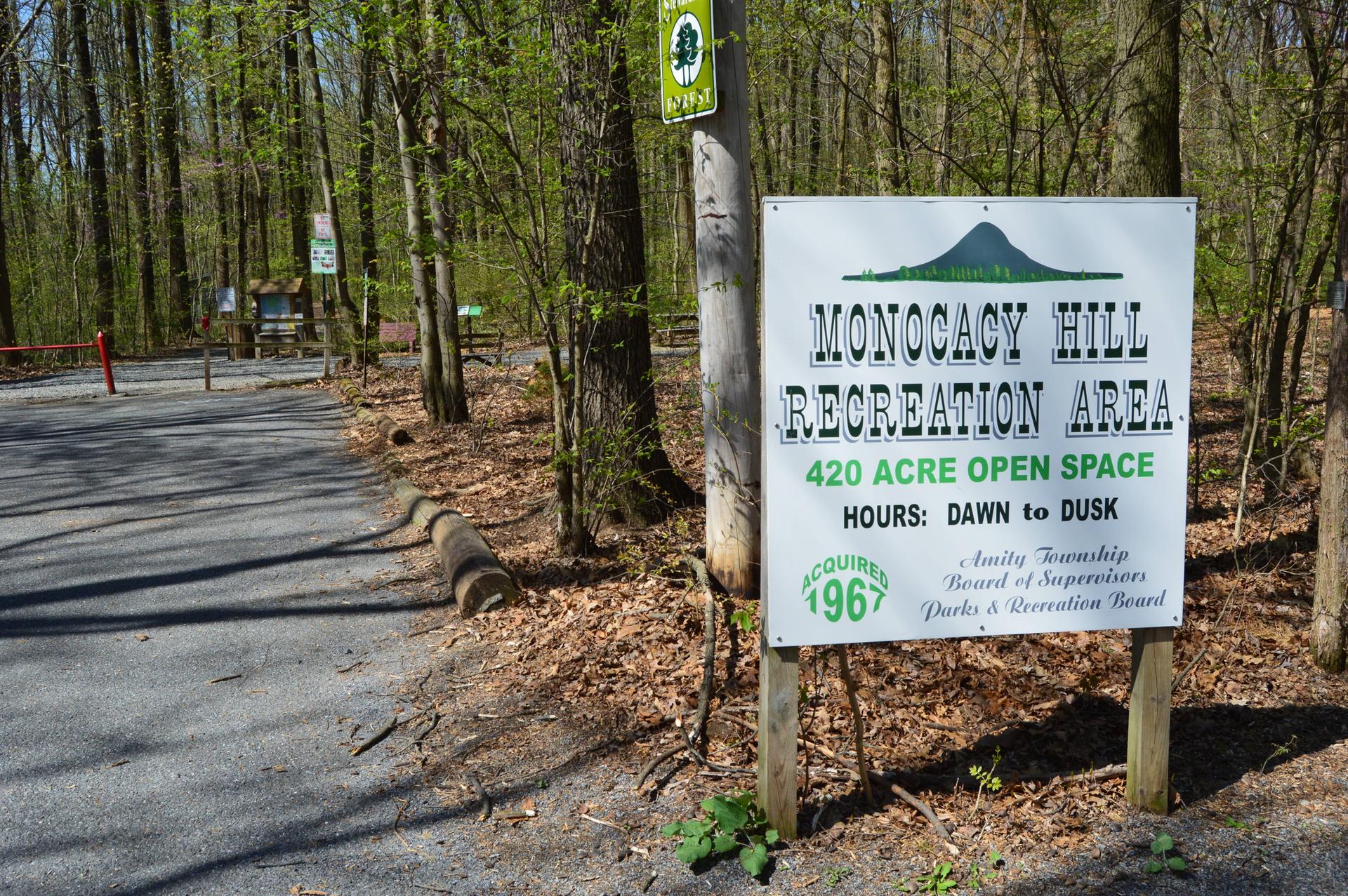 A white sign stands beside a paved path at a sunny forest entrance.