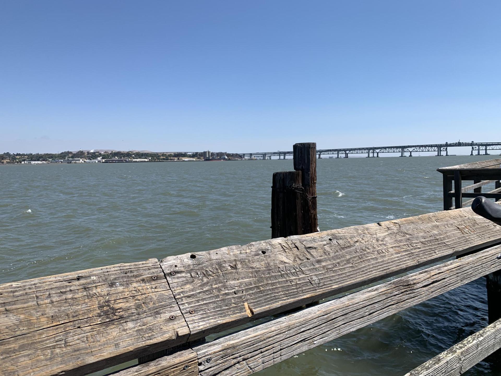 Wooden pier overlooking a bay with a distant bridge on a sunny day.