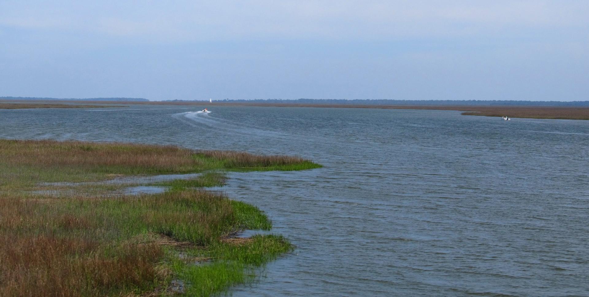A boat moves across wide water with marsh grass under an overcast sky.
