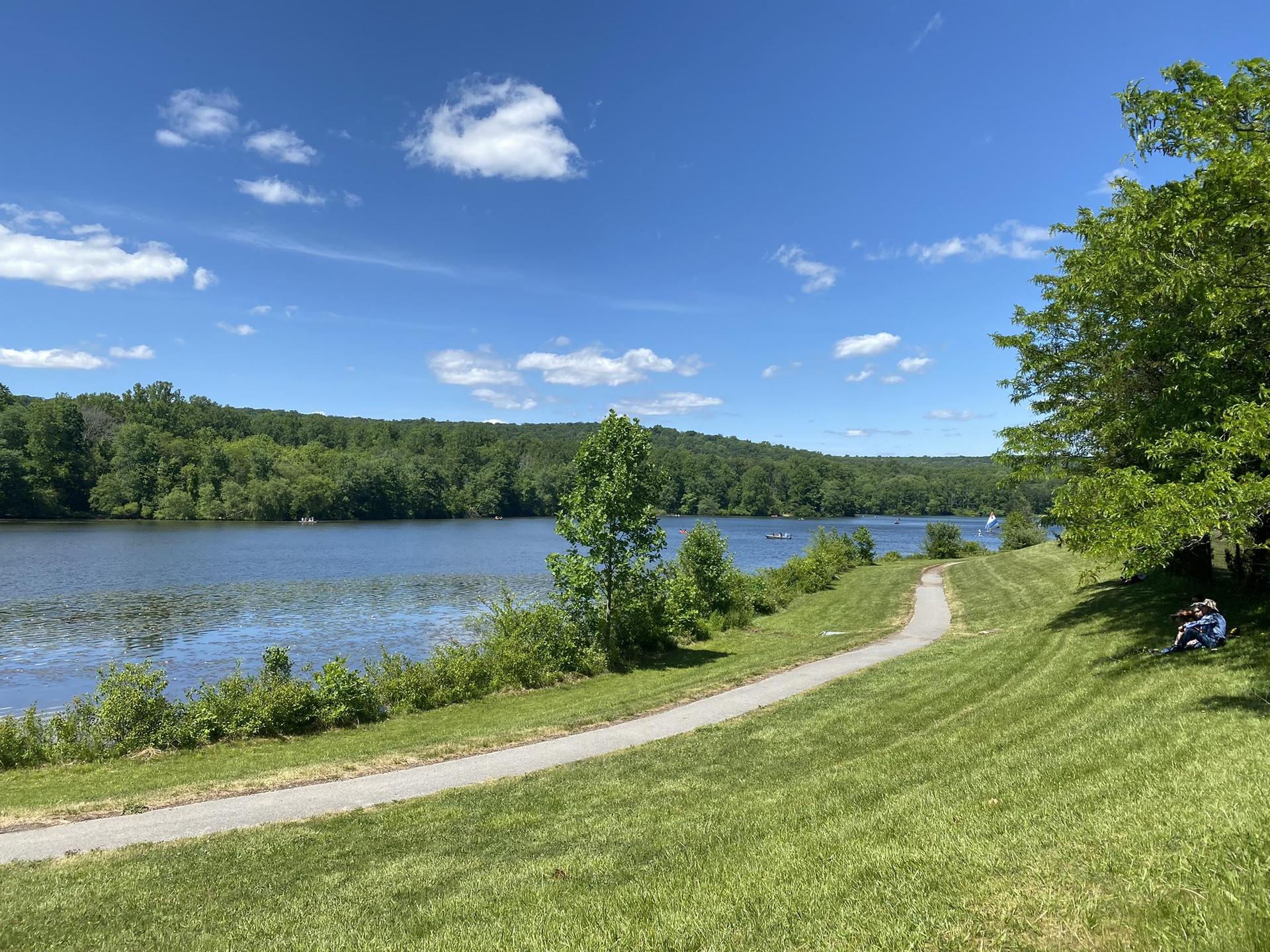 Sunny lake view with a paved path, green hills, and people relaxing.