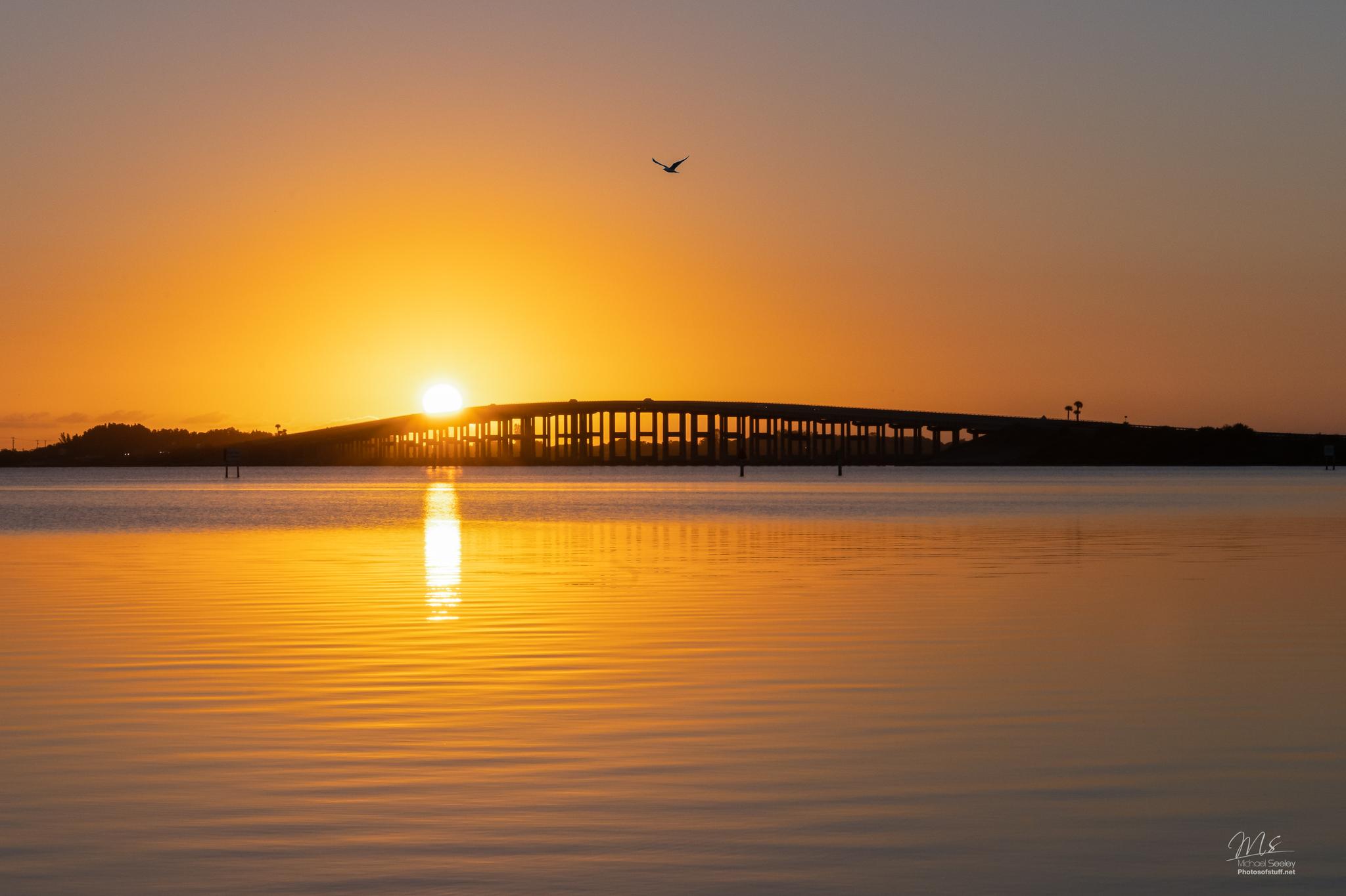 A long bridge is silhouetted against a vibrant golden sunset over calm water.