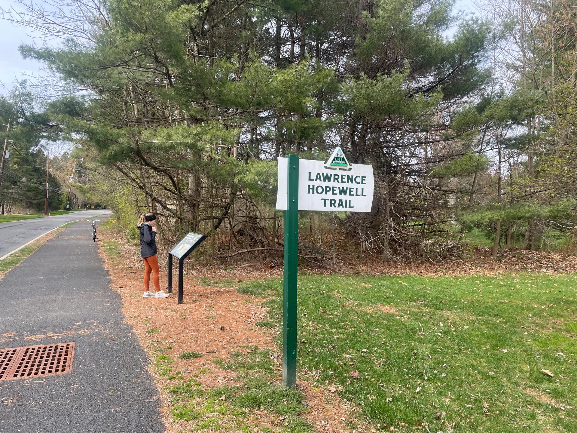 Person looking at a trail sign next to a paved path and pine trees.