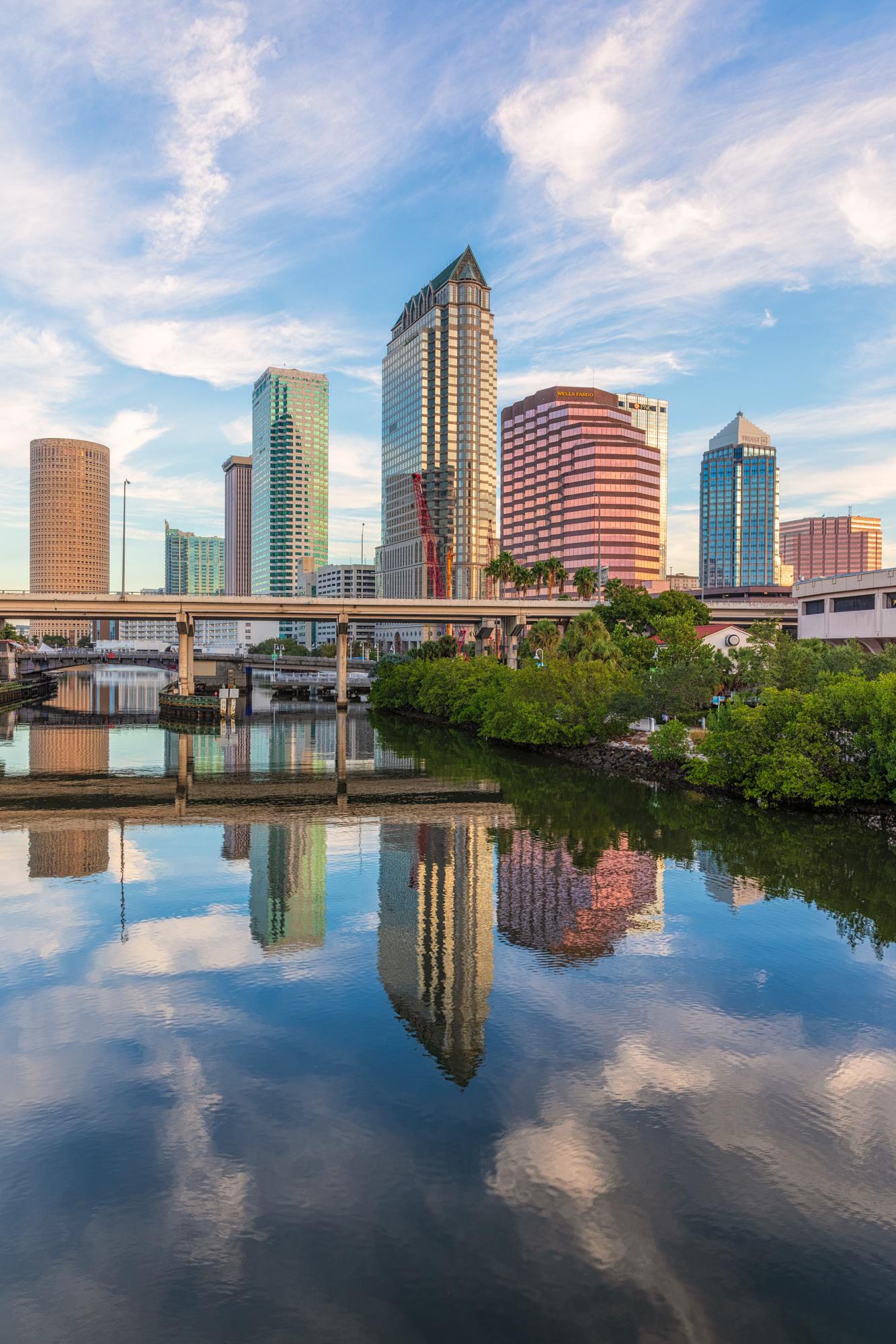 City skyline reflecting in calm water, with a bridge and bright sky.
