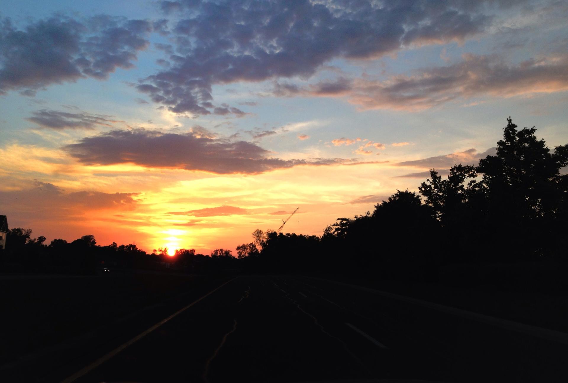 A vibrant sunset over a dark road, silhouetting trees against a colorful sky.