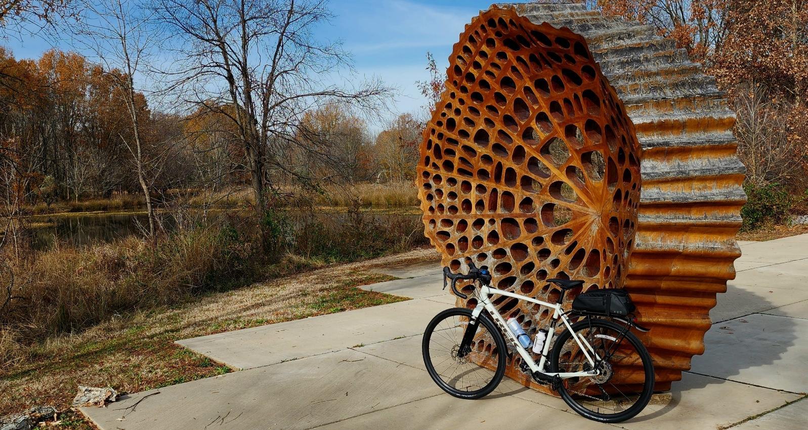 A bicycle rests beside a large rusty metal sculpture on a paved path.